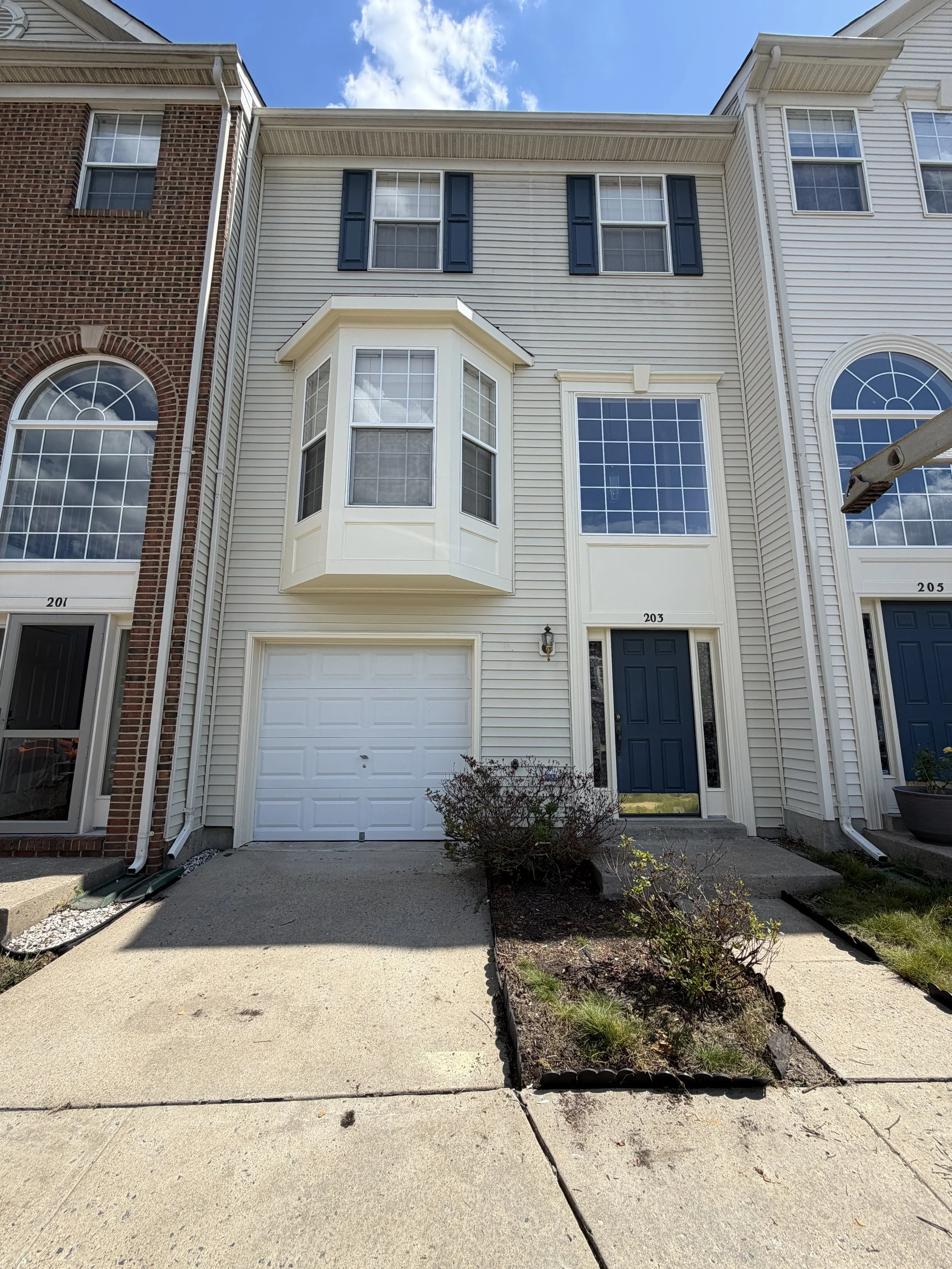 View of three attached townhouses with bay windows and front doors, one has a garage, and a small garden bed with plants in front.