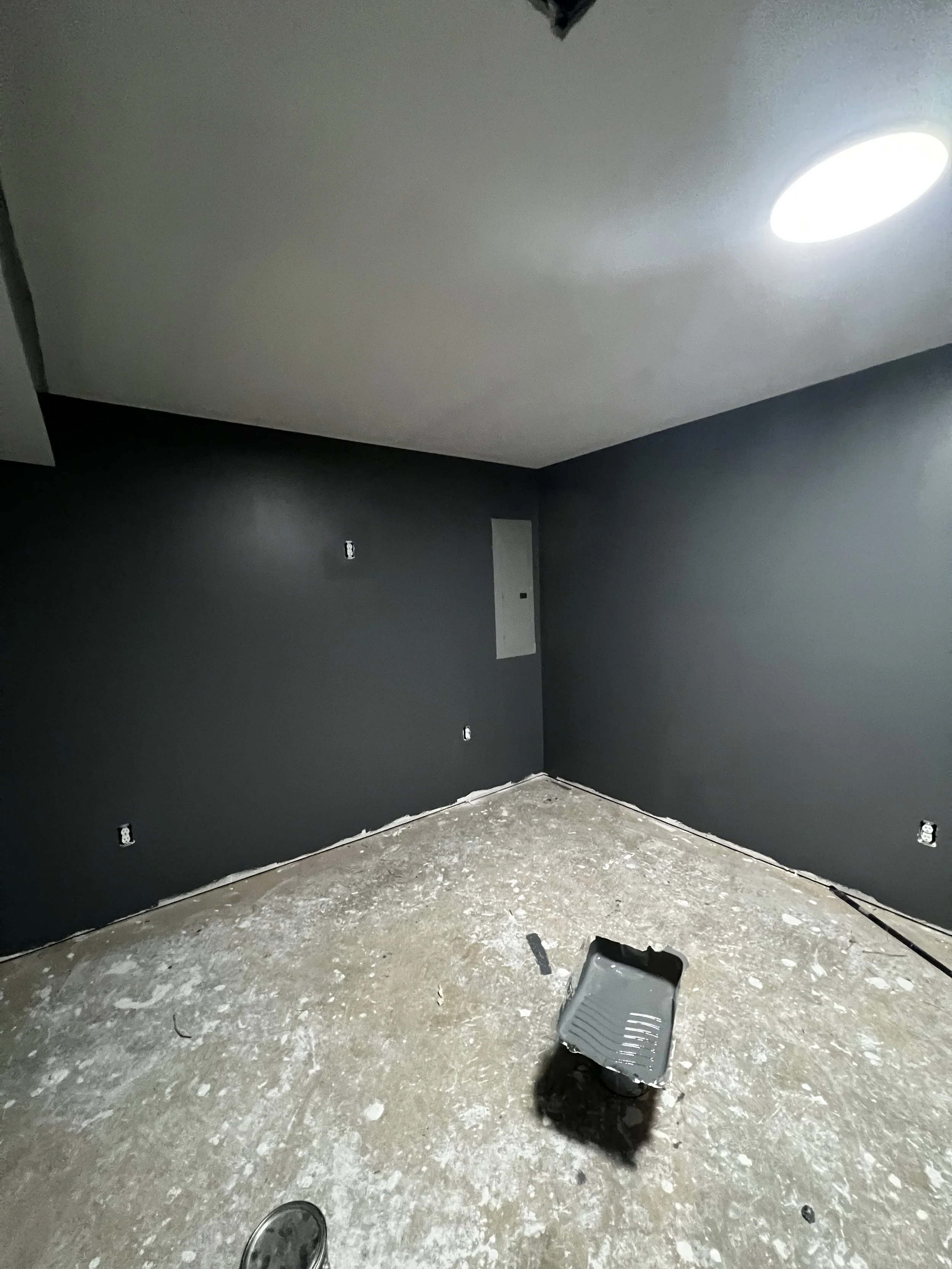 unfinished room with dark gray walls, exposed electrical outlets, an open gray plastic container on the floor, and a single bright ceiling light. The floor is covered in debris and dust.