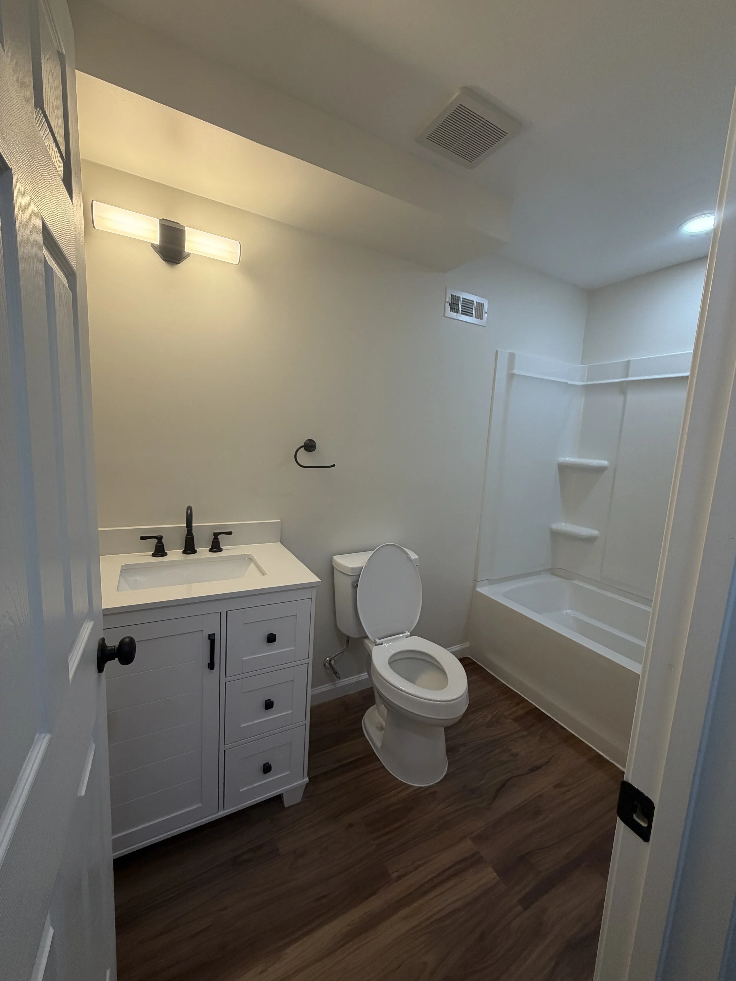 A small bathroom with a white vanity, a mirror with a light fixture above it, a toilet, and a shower with a built-in bench and a ceiling vent, all on wood flooring.