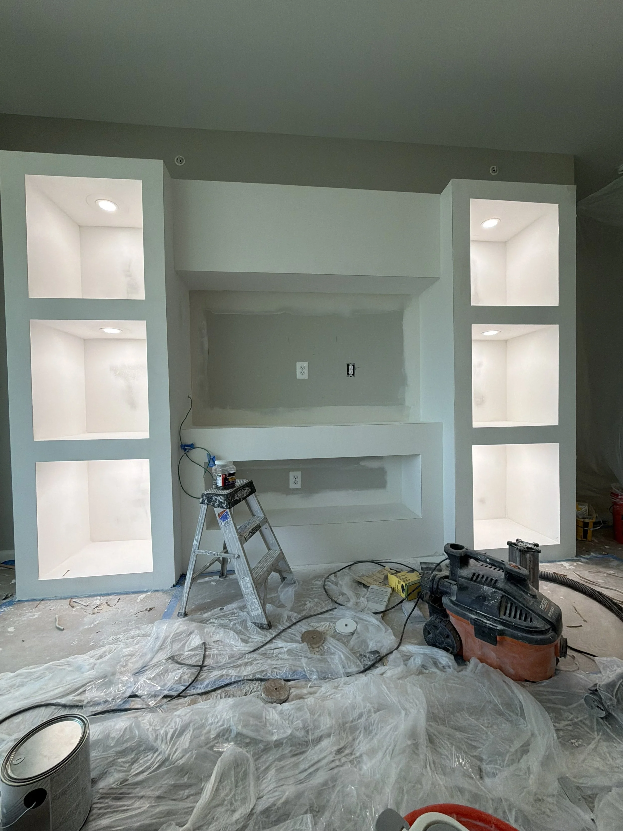 A room under construction with built-in white shelves, a ladder, tools, and paint supplies scattered on the floor covered with plastic sheeting.