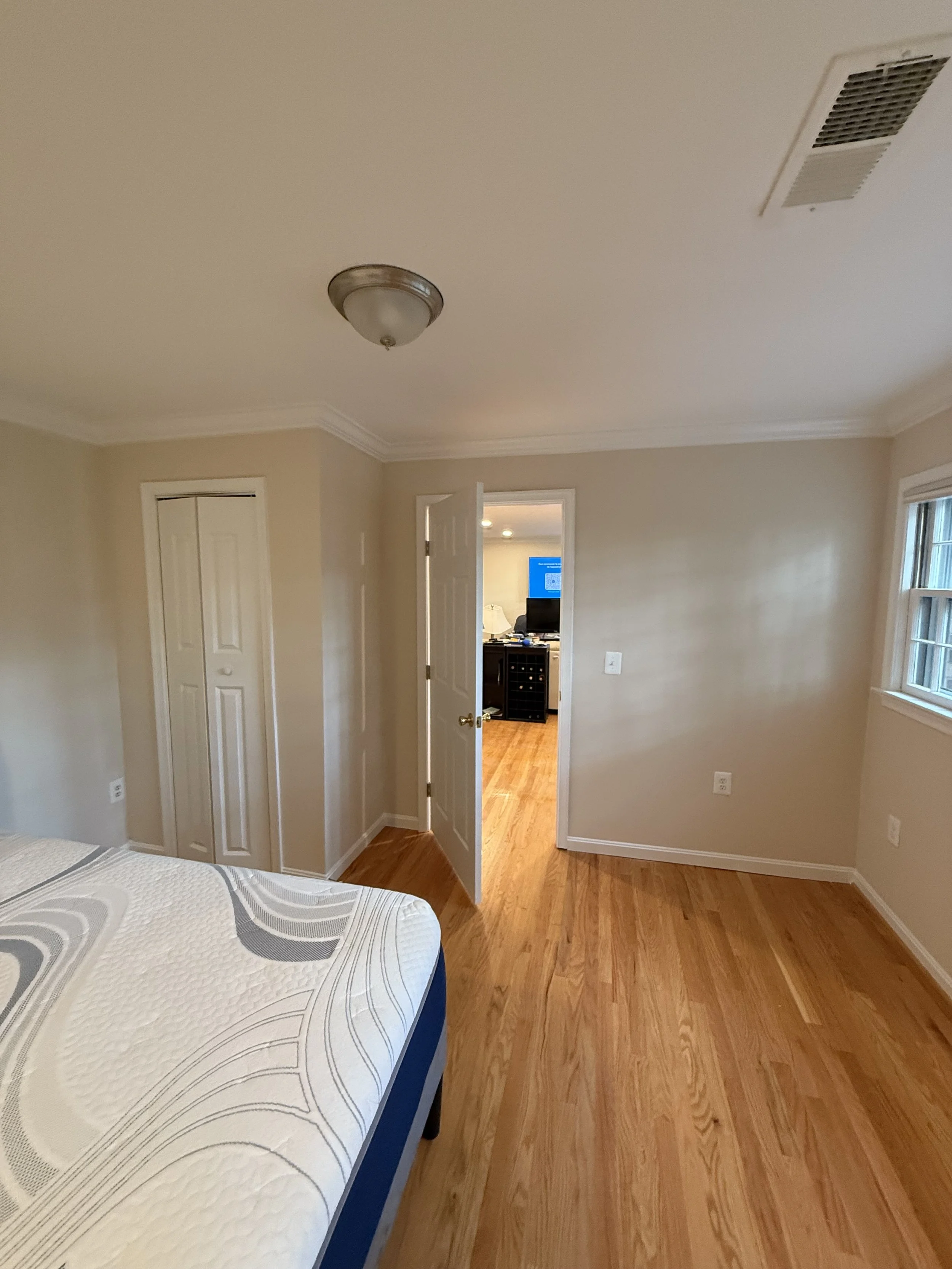 A bedroom with beige walls, hardwood floor, a bed with a white and gray patterned mattress, a closet with sliding doors, and an open door leading to another room with a TV and a small cabinet.