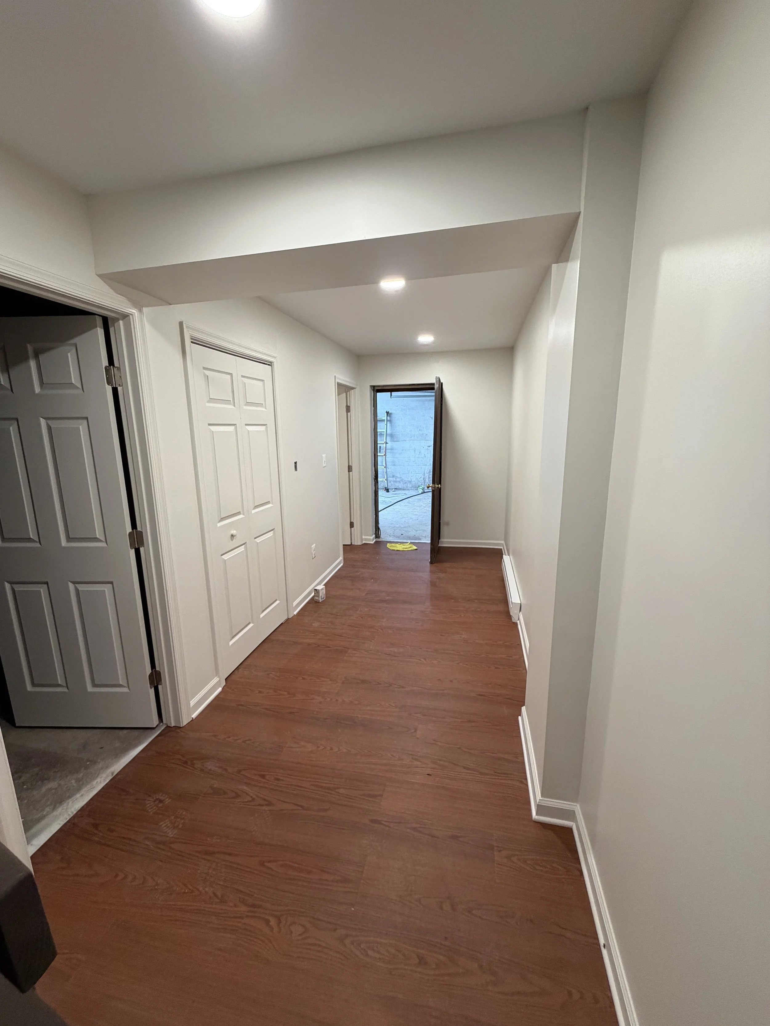 Empty hallway with white walls, hardwood floors, and a door open to an outdoor area with a concrete wall and ladder.