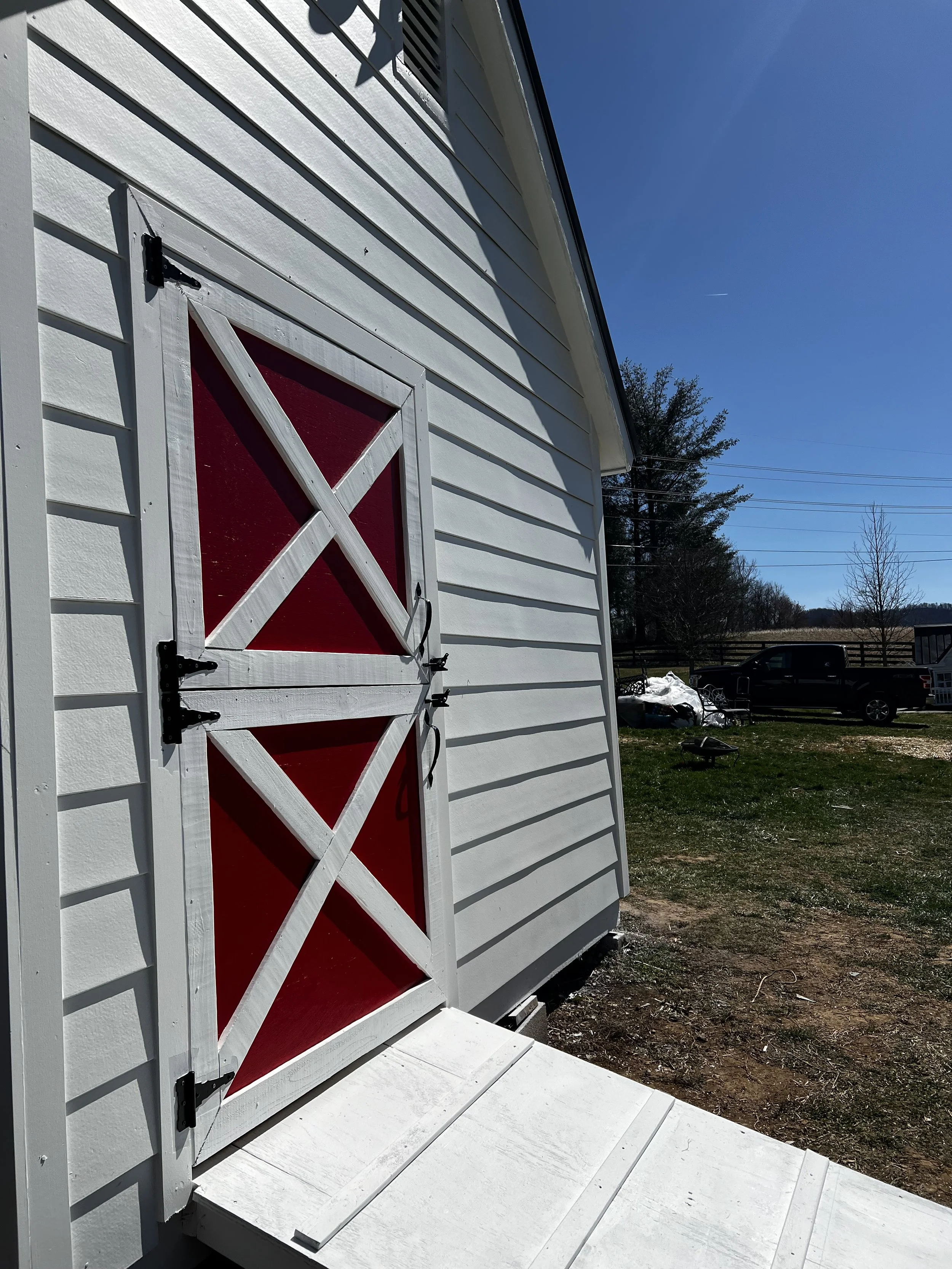 Close-up of a white barn door with red accents, mounted on a white house with horizontal siding, with a background of a grassy yard, trees, a black truck, and a clear blue sky.