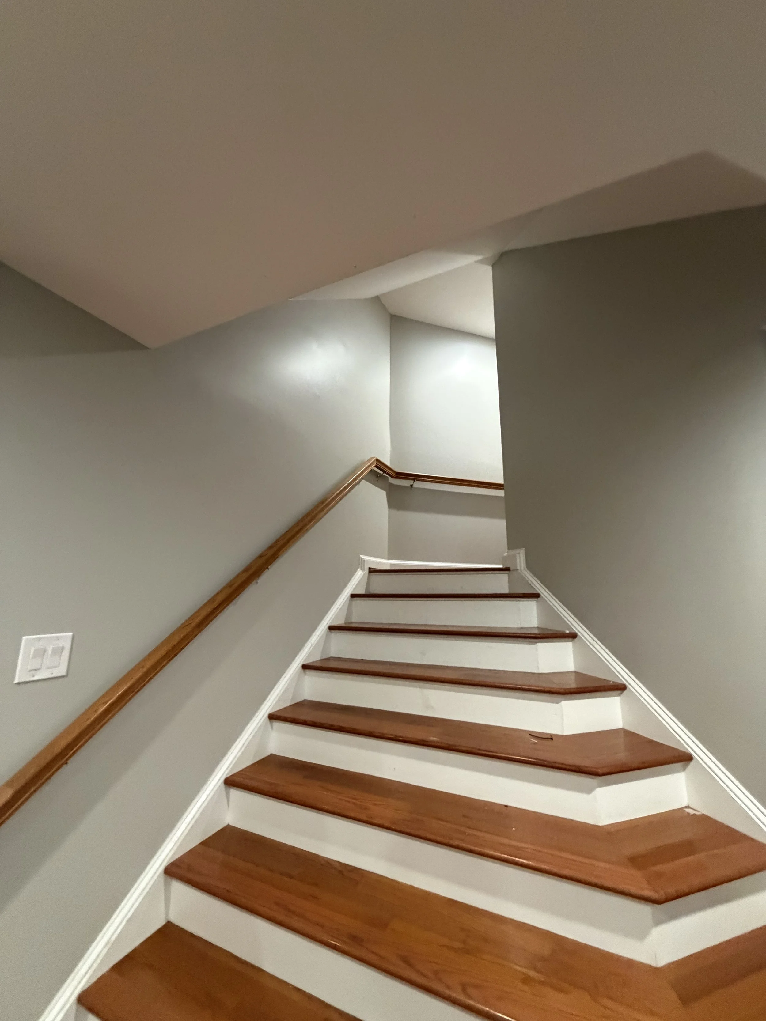 A staircase with wooden steps and a wooden handrail, leading upward in a residential interior with gray walls and white trim.