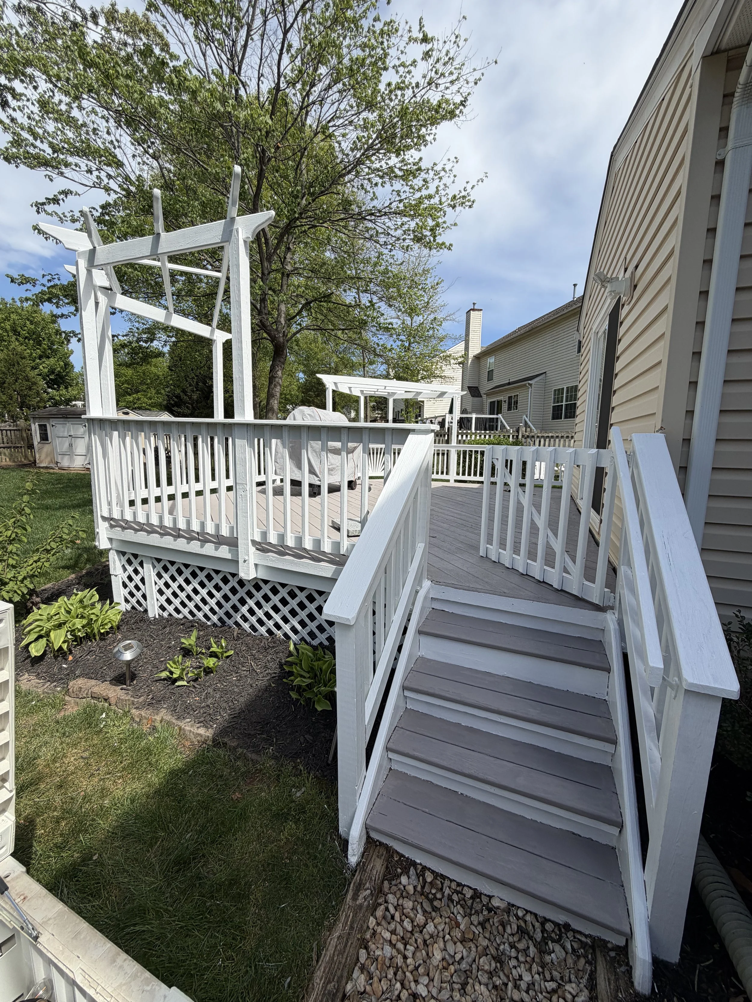 A white wooden deck with stairs leading down to a garden with green grass and plants, a yellow house with beige siding, and a tree with green leaves in the background.