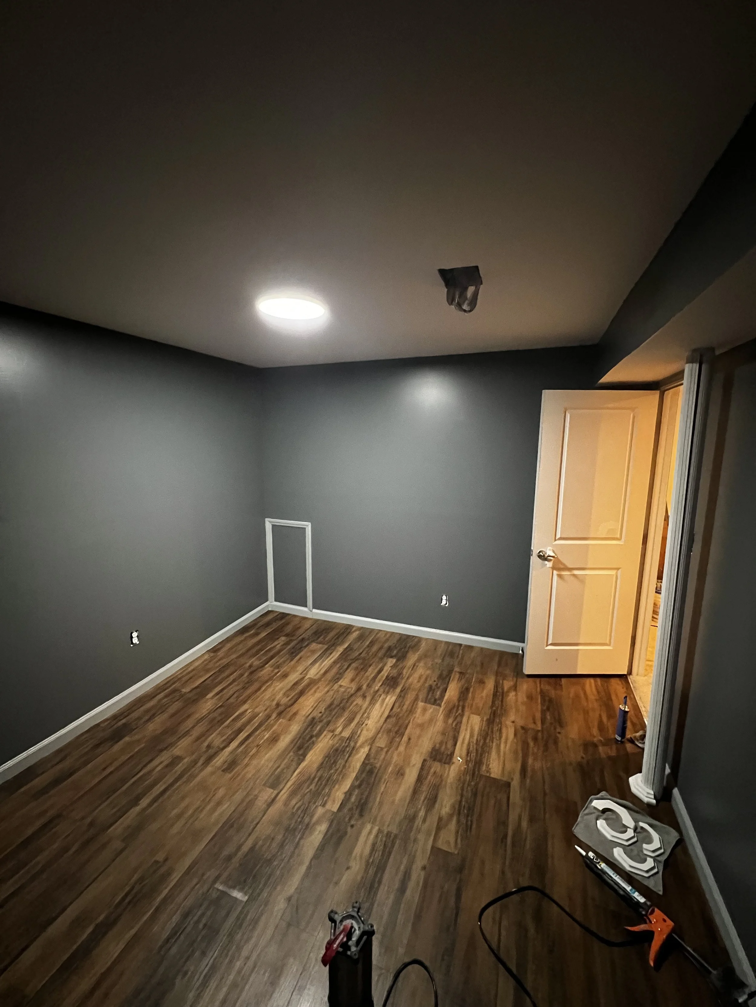Room with dark grey walls, hardwood flooring, ceiling light, partially open door, construction tools, and materials on the floor, indicating ongoing renovation or flooring installation.