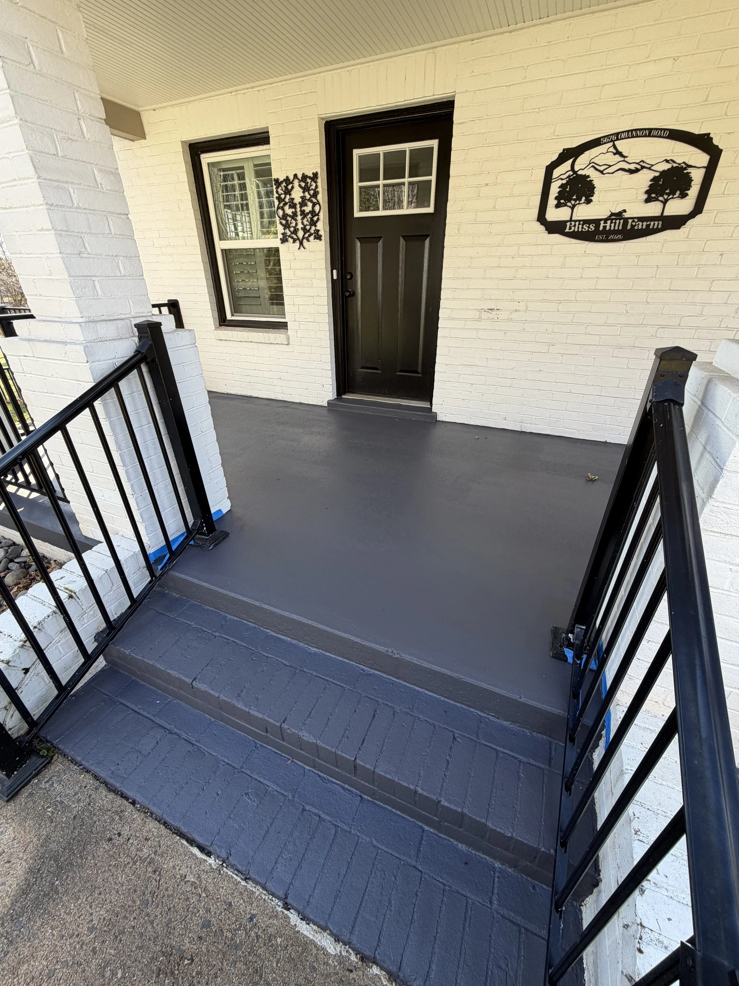 Front porch of a house with black door, white brick wall, window with shutters, decorative metal wall art, and a black metal railing on stairs, with a wooded area visible in the reflection.