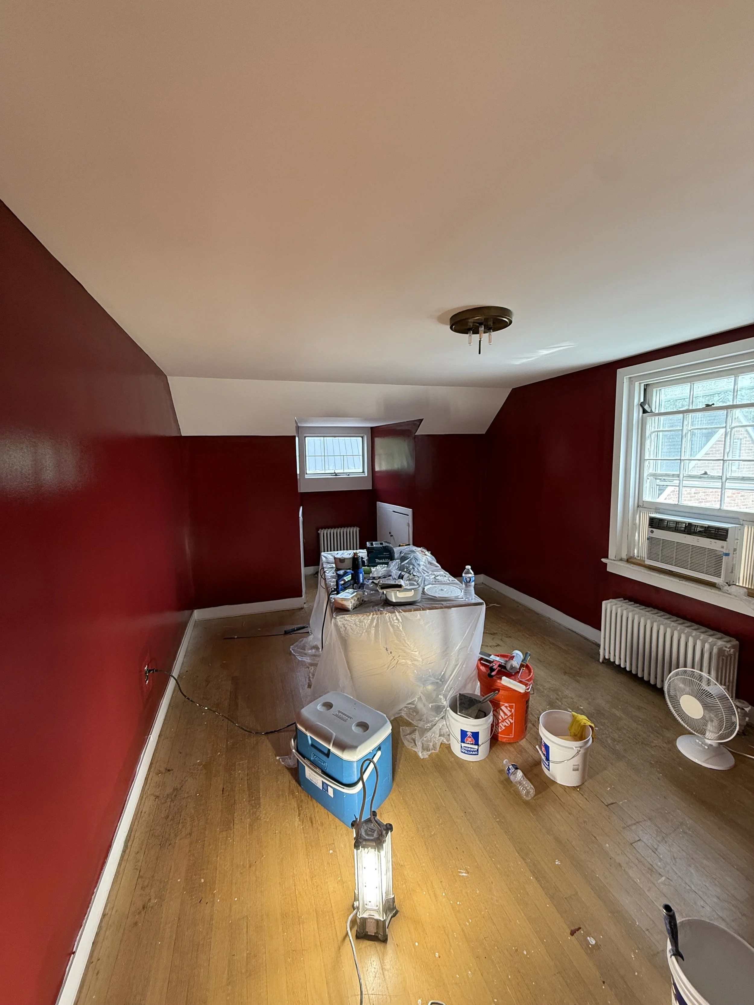 A room under renovation with red walls, a window air conditioning unit, a small window, and painting supplies on the floor, partially covered by a plastic sheet, with an extension ladder, buckets, and tools scattered around.
