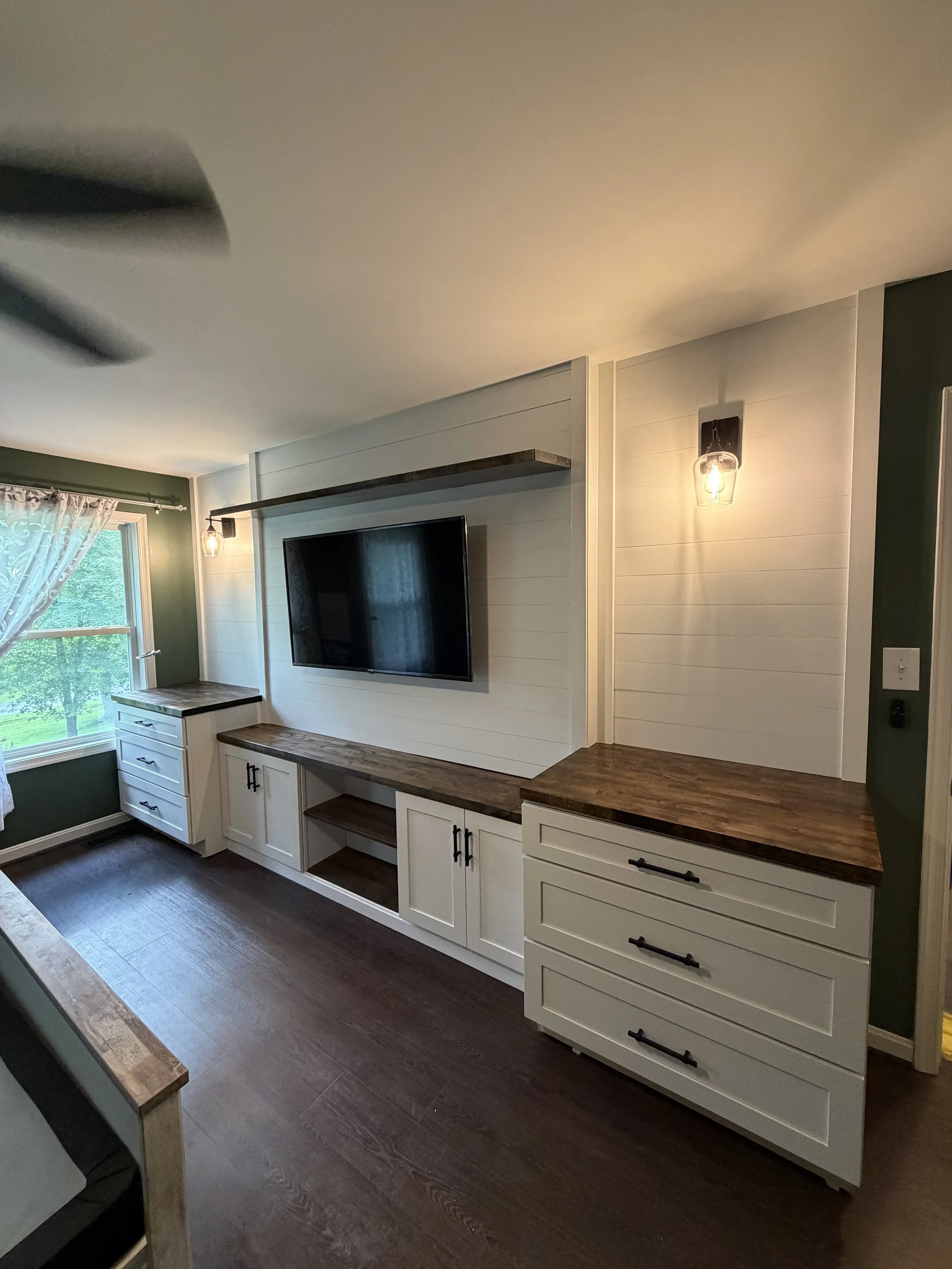 Interior view of a living room with a wall-mounted television, white cabinetry with dark wood countertops, a window with curtains, and a dark wood floor.