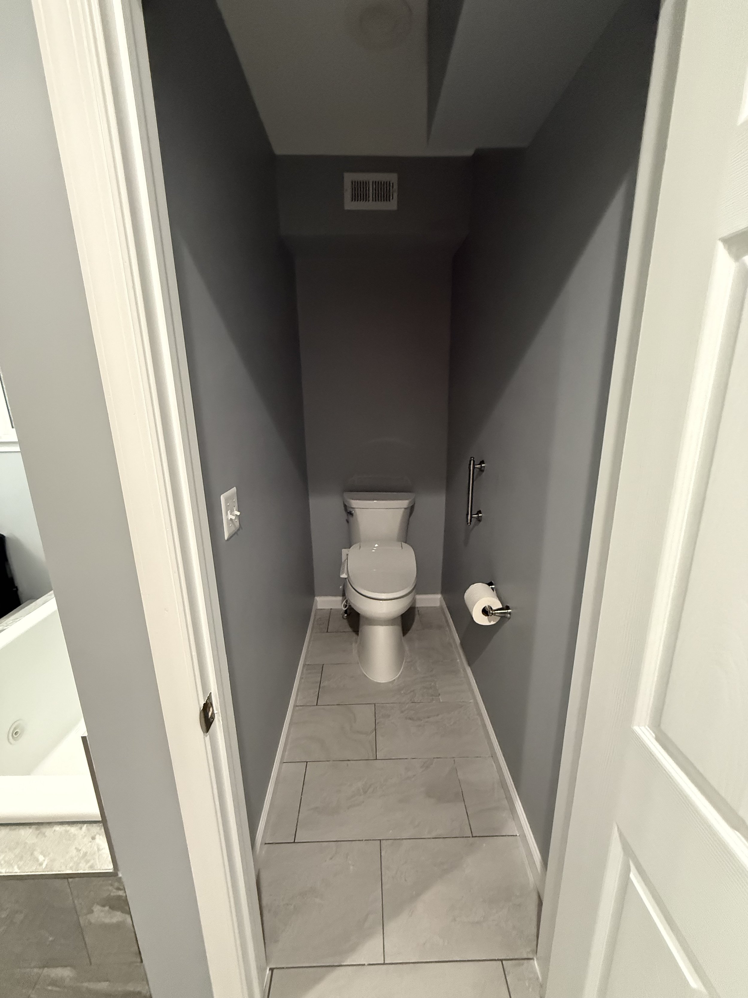 A small bathroom with gray walls and gray floor tiles, containing a white toilet, a wall-mounted handrail, a vent on the ceiling, and a light switch on the wall.