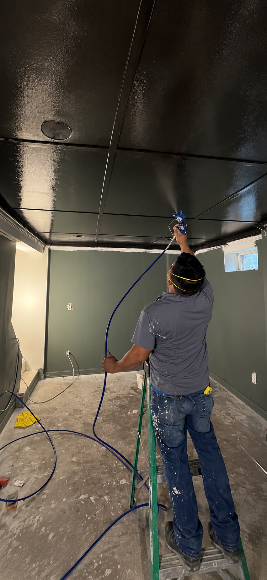 A man standing on a step ladder, spray painting a black ceiling in a room. He is wearing a gray shirt, jeans, and safety goggles, with paint splatters on his clothes and a yellow cloth on the floor nearby.