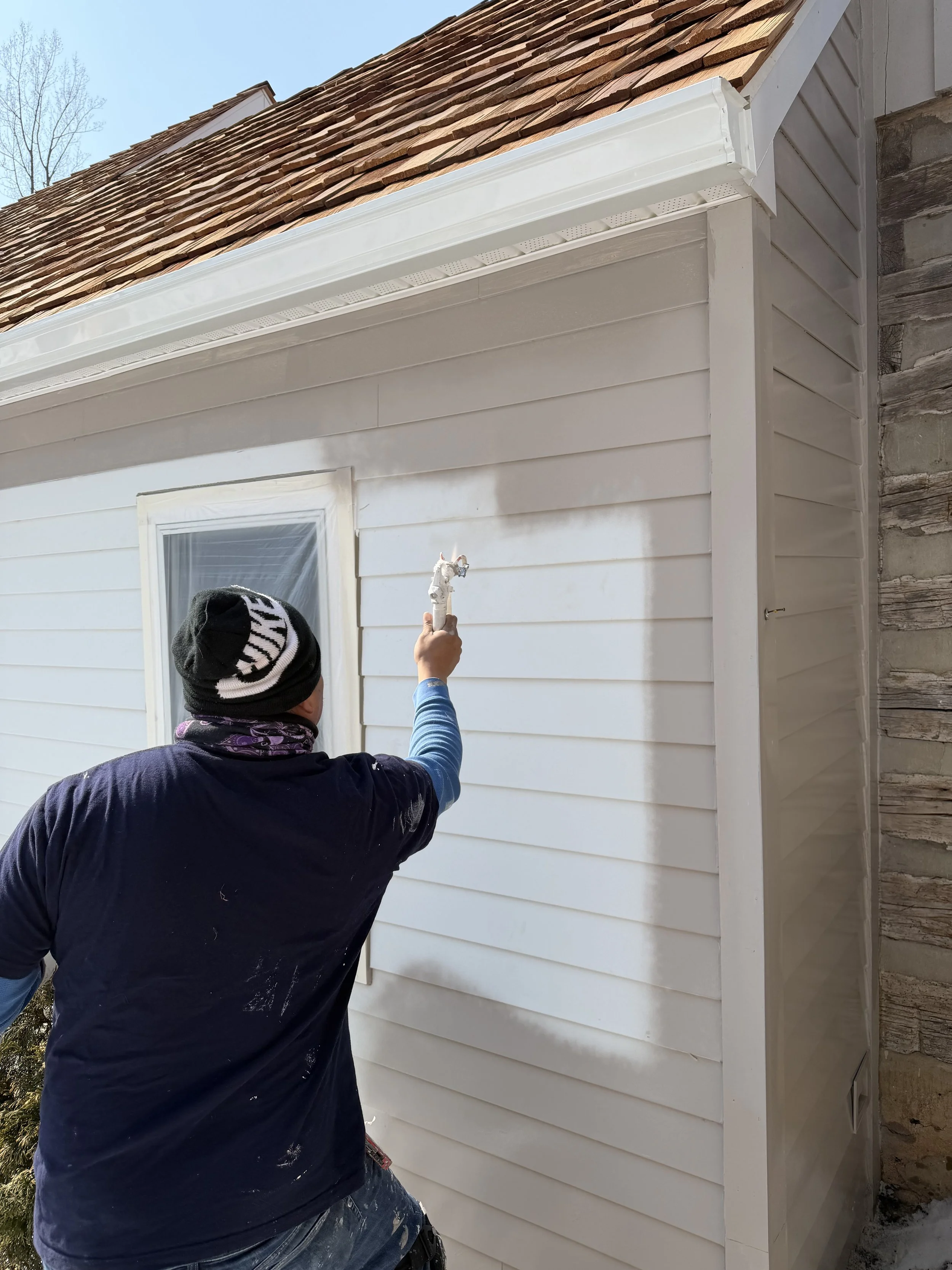 A person painting the exterior wall of a house white using a paintbrush. The house has horizontal siding and a window with a white frame. The person is wearing a black jacket, a beanie, and gloves, and is working in daytime with a clear sky.