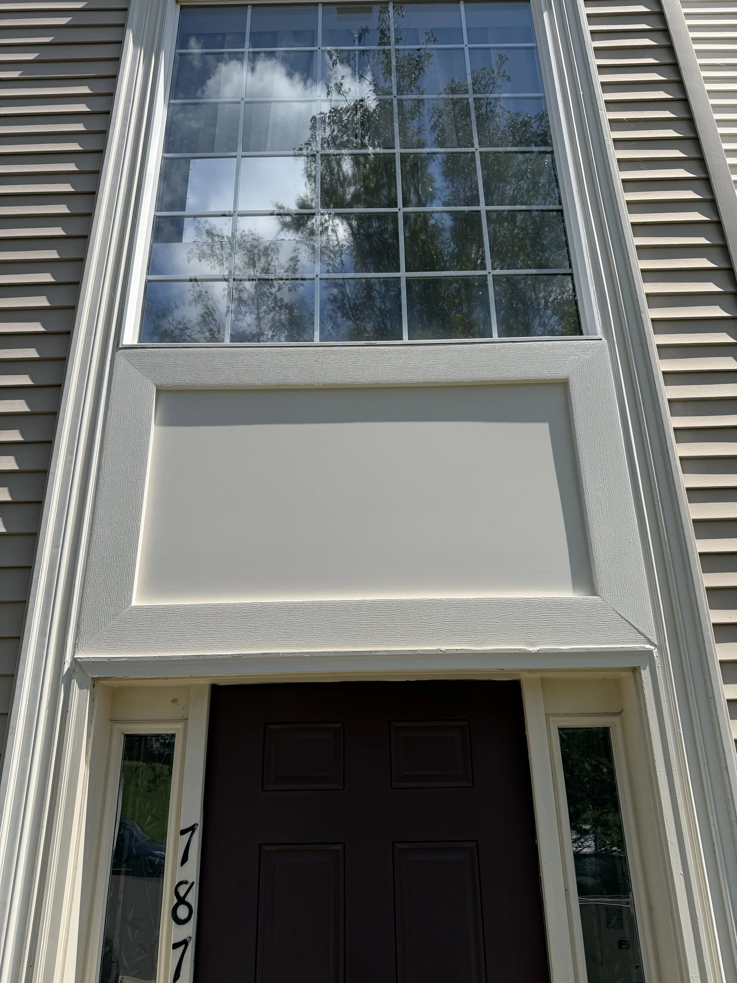 Front door of a house with a large window above, showing reflection of a cloudy sky and trees. The house has beige vinyl siding, white trim, and house number 718 on the left side of the door.