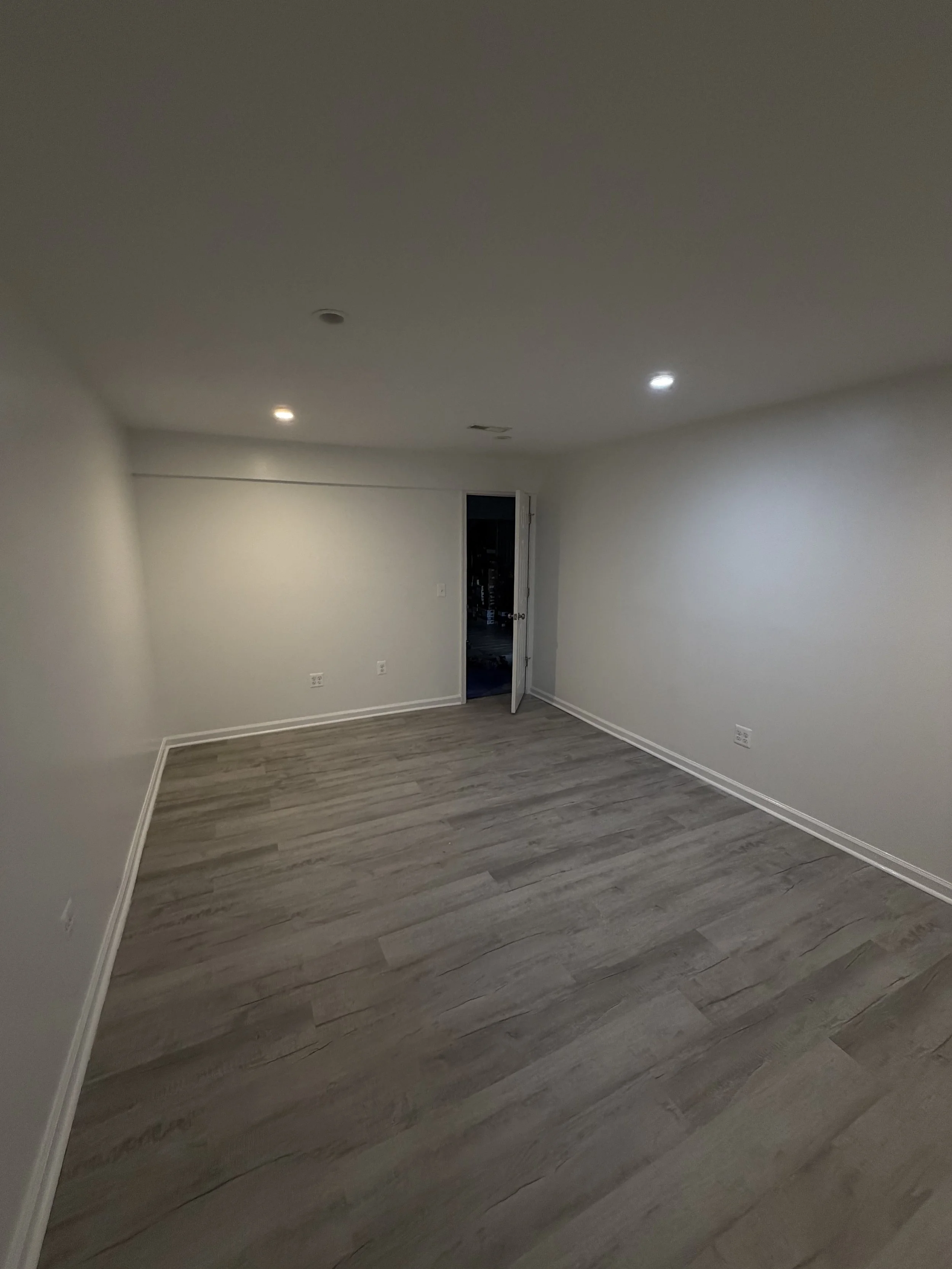 An empty room with white walls, gray wood flooring, and a partially open door leading to a dark room.