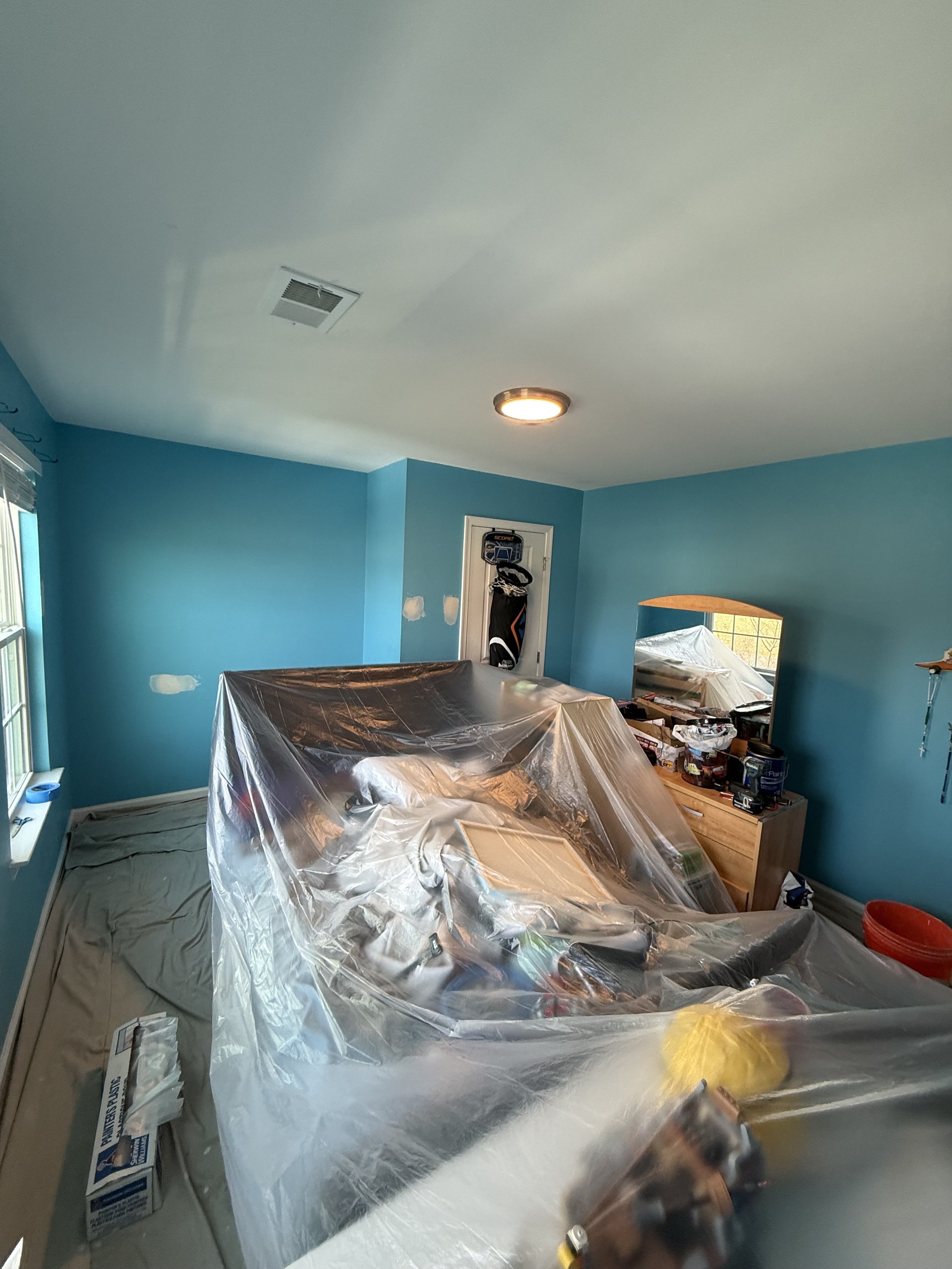 Room undergoing renovation with walls painted light blue, covered furniture, and painting supplies on a wooden dresser.