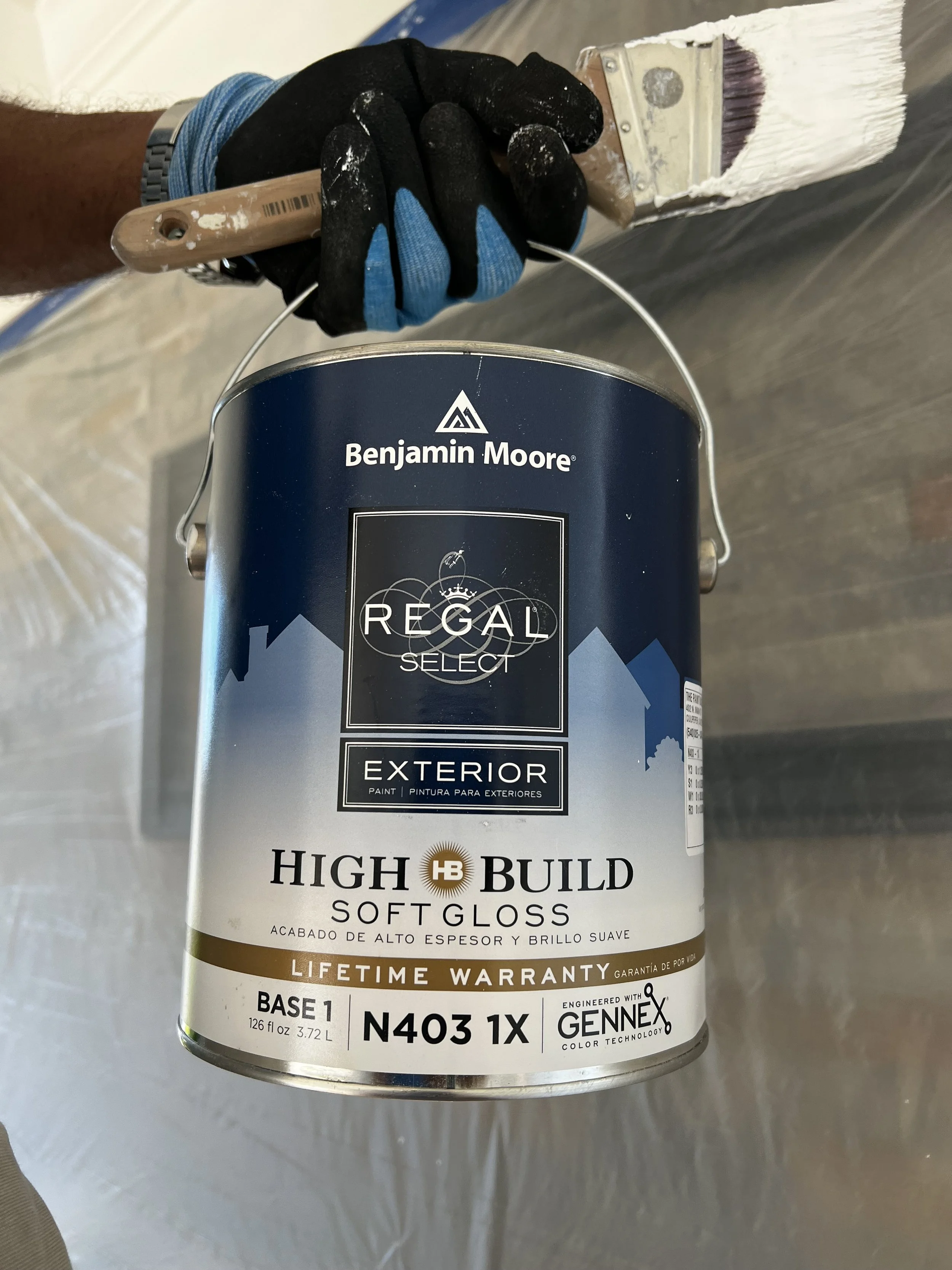 A person holding a can of Benjamin Moore Regal Select exterior soft gloss paint, wearing a black glove and a silver watch, with part of their arm and clothing visible.