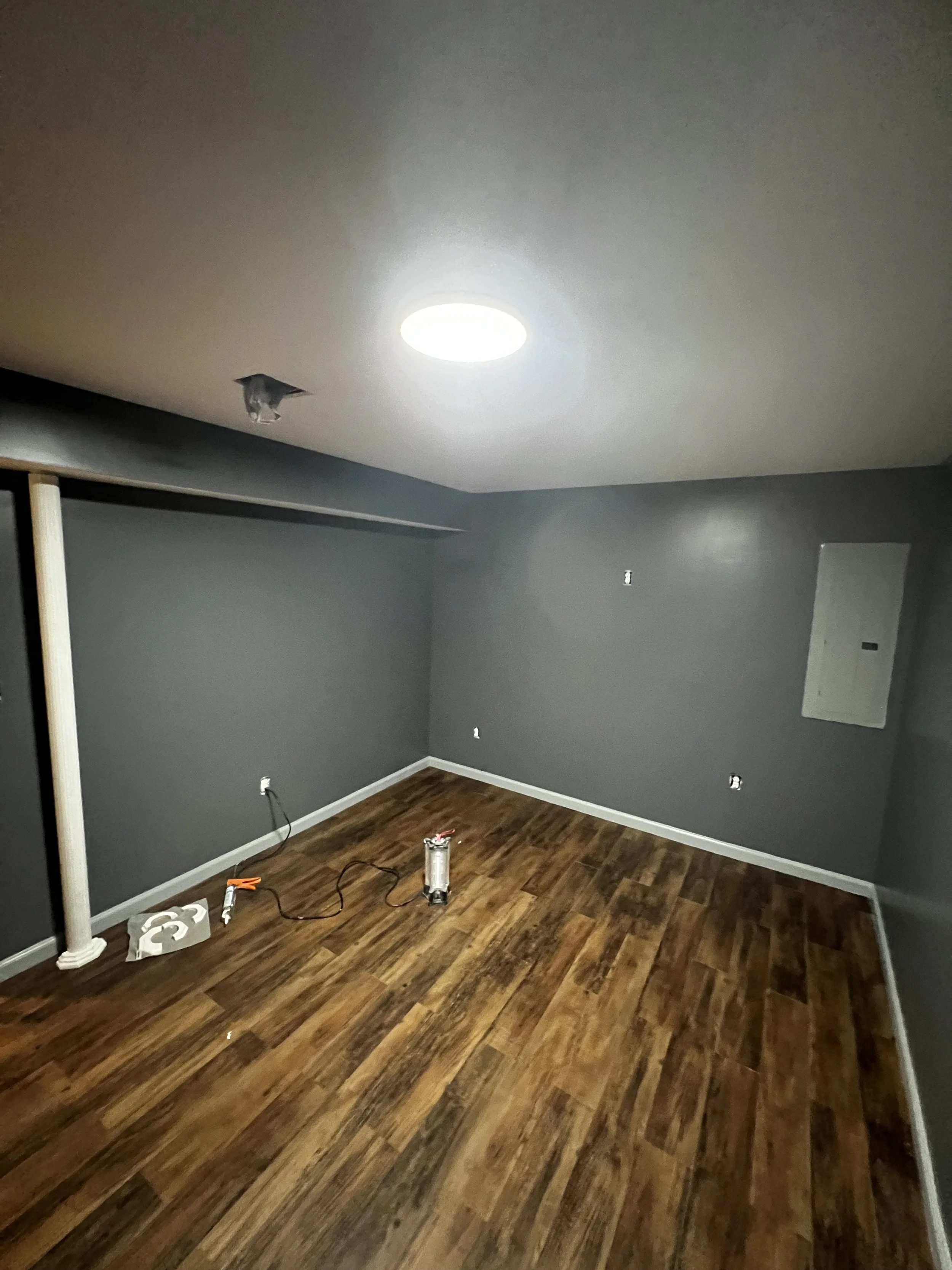 Empty room with dark gray walls and a wooden floor. The ceiling has a bright round light fixture and a small hole with a piece of fabric hanging out. On the floor, there are some tools and a small metal canister. An electrical cord is also present.