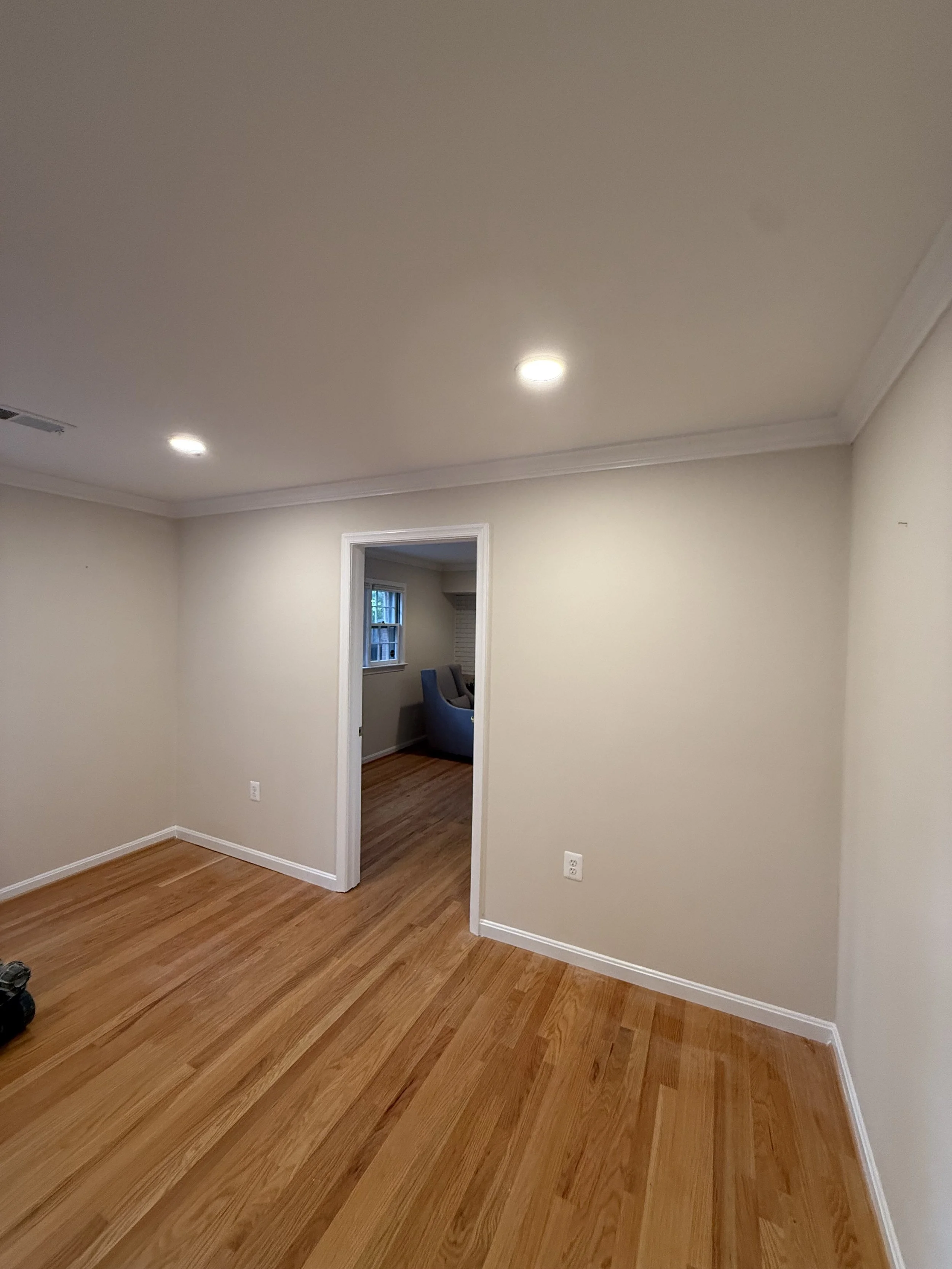 Empty room with beige walls, hardwood floors, white trim, and a doorway leading to another room with a window and chairs.