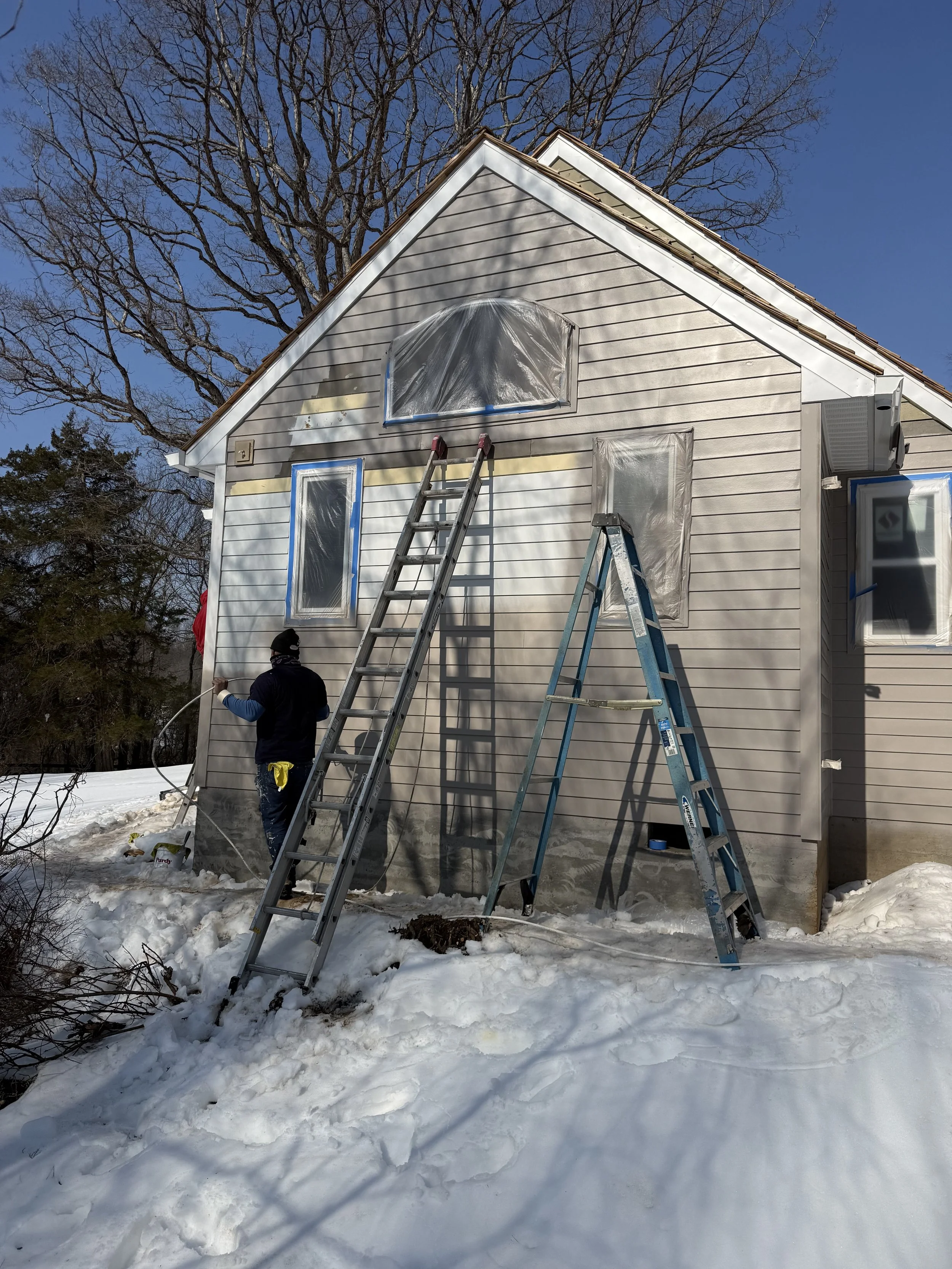 A person is painting the exterior wall of a house in winter, with ladders leaning against the house, plastic covers over windows, and snow on the ground.