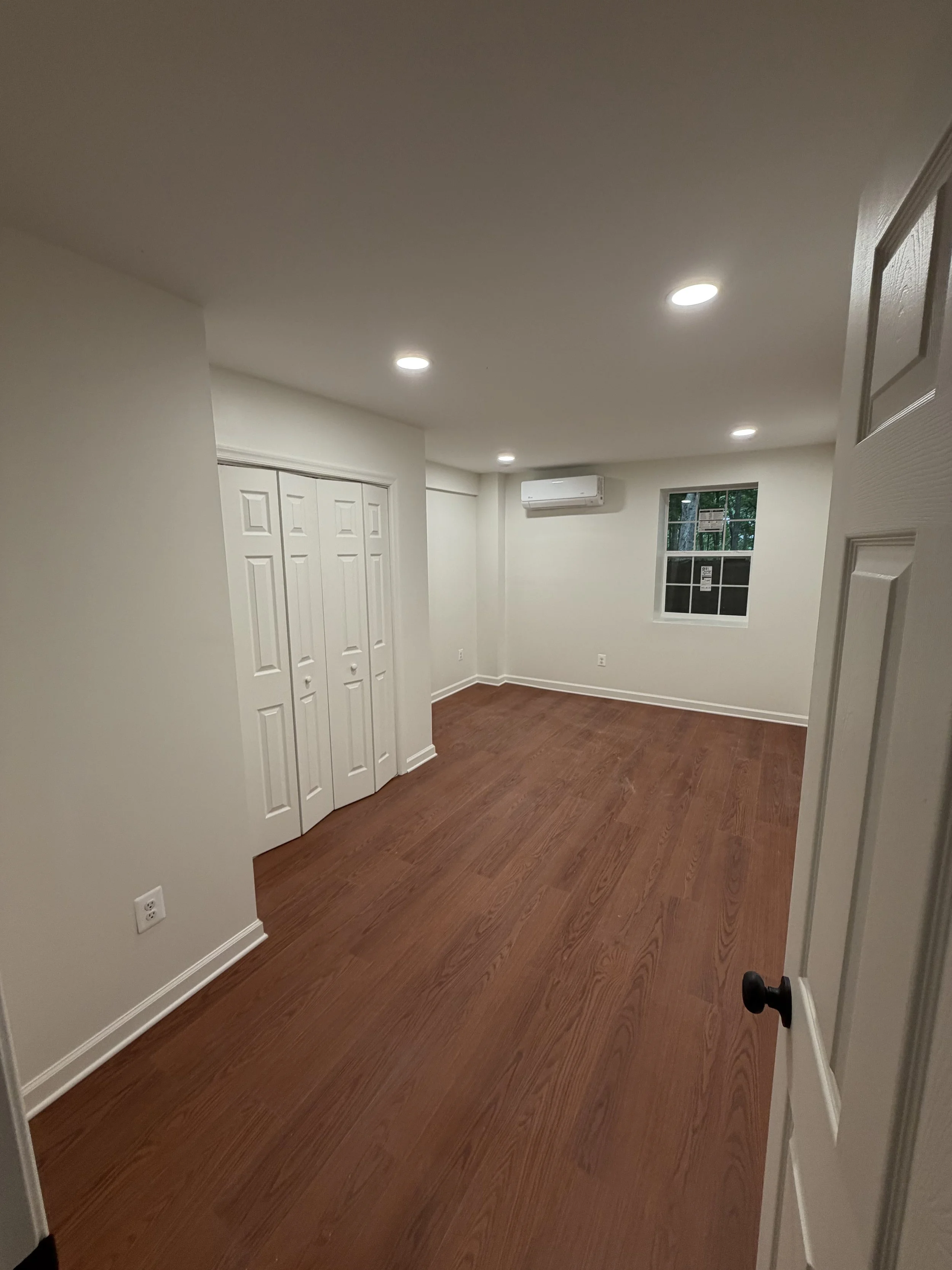 Empty room with hardwood floors, white walls, ceiling lights, a window, an air conditioner, and a door open to this room.