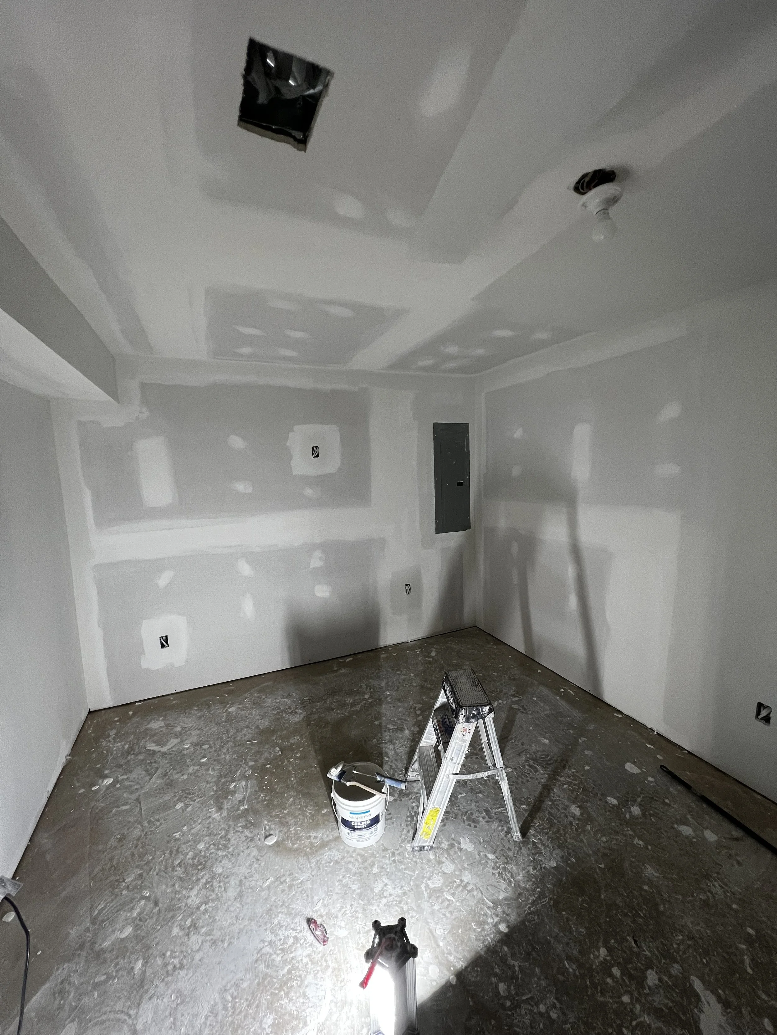 A room under construction with drywall walls and ceiling, a small ladder, a work light on the floor, and painting or renovation supplies, including a bucket of primer or paint.