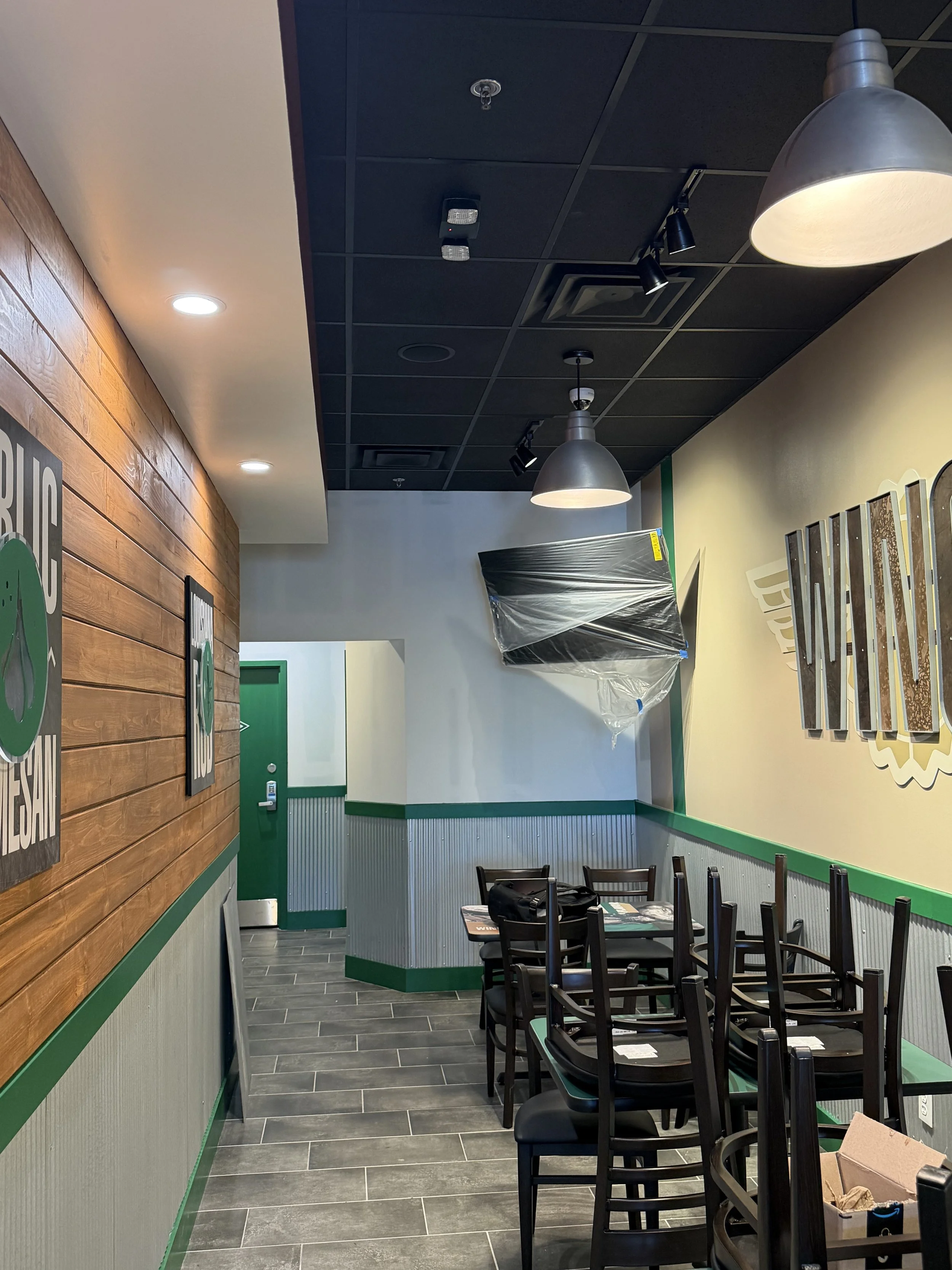 Interior of a restaurant or café with black chairs, a wooden wall with several signs, a painted wall with decorative elements, and a table in the foreground with a box and some papers. The ceiling has black panels, light fixtures, and track lighting.