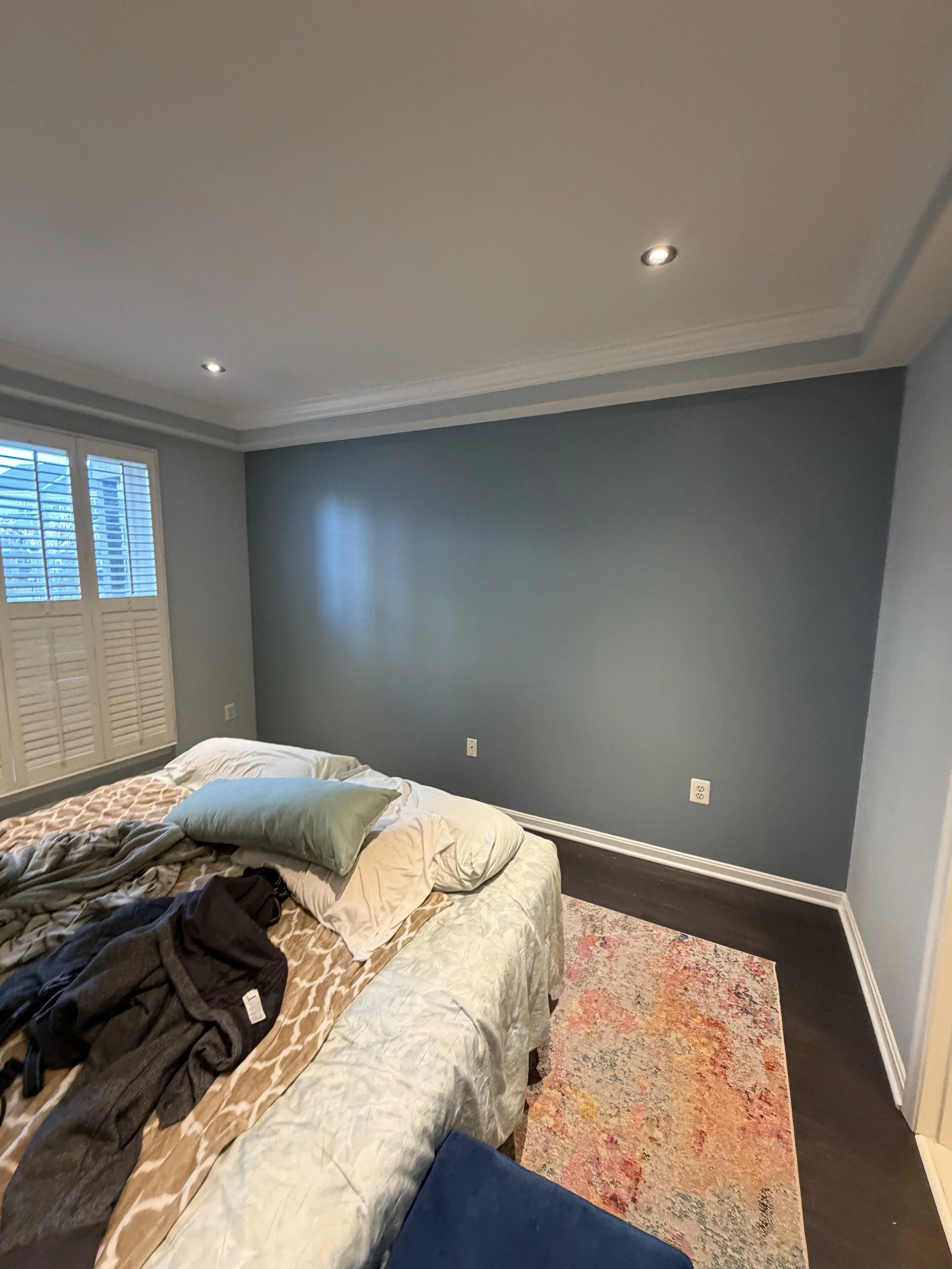 A bedroom with a bed covered in a beige and brown patterned blanket, messy with clothing and pillows. The wall behind the bed is painted blue, with multiple electrical outlets. There is a window with white shutters and a floral area rug on dark hardw