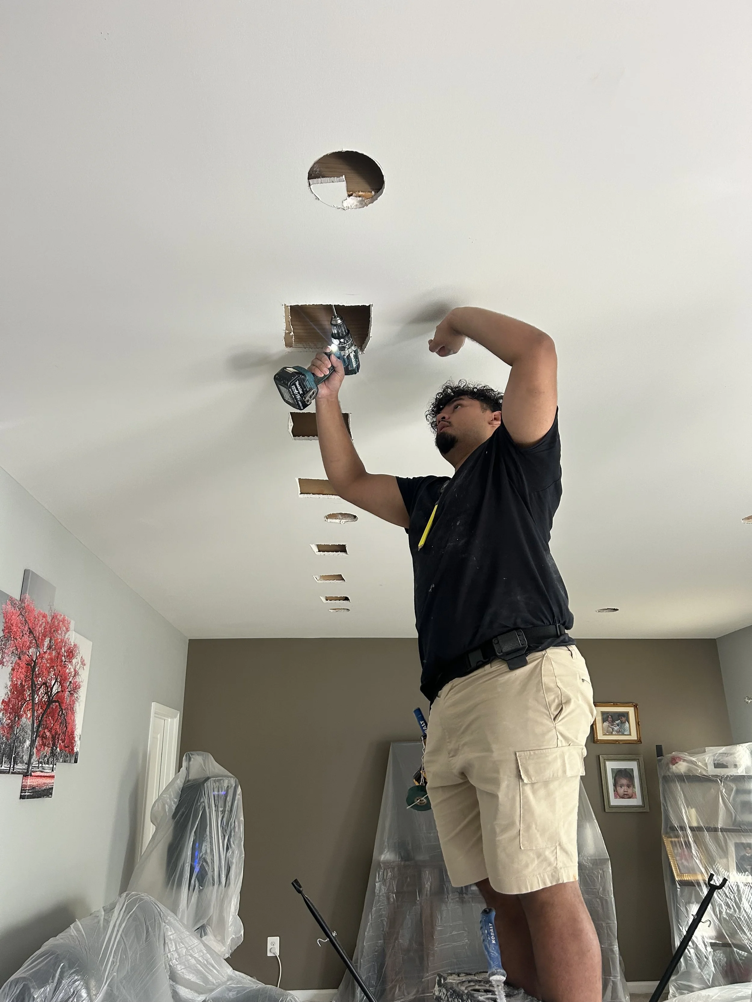 Man installing or repairing a ceiling light fixture with a drill, in a room with wall art, framed photos, and covered furniture.