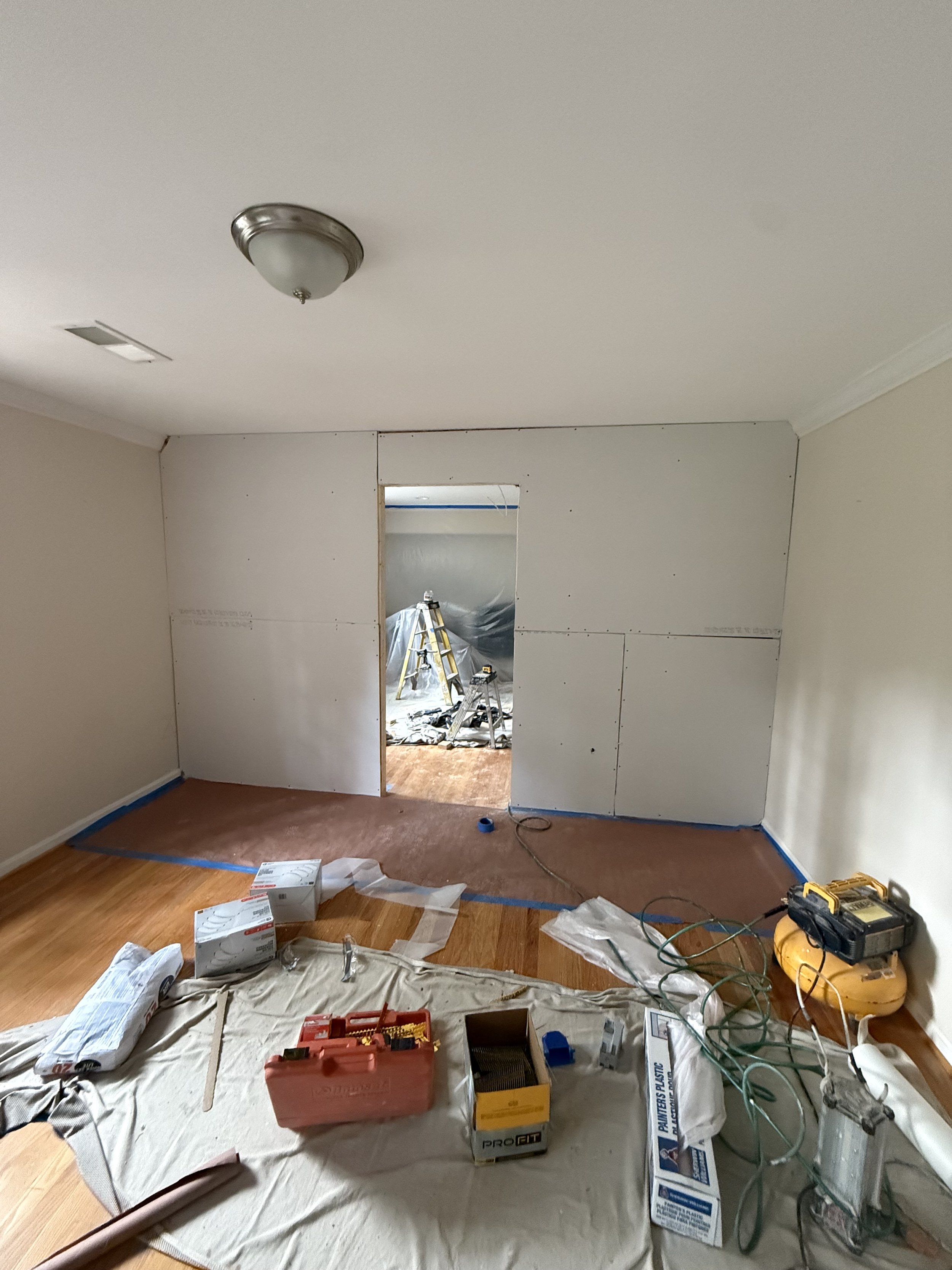 Room under renovation with drywall being installed, tools, supplies, and materials scattered on a protective sheet on the floor.