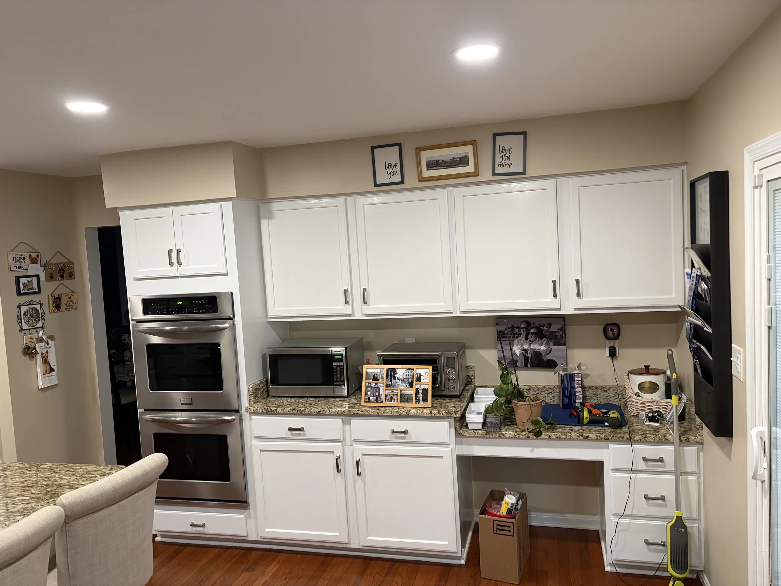 Kitchen with white cabinets, granite countertops, stainless steel appliances, and decorative wall art including framed photos and quotes, some artwork leaning on the counter, and a dining area with beige upholstered chairs in the foreground.