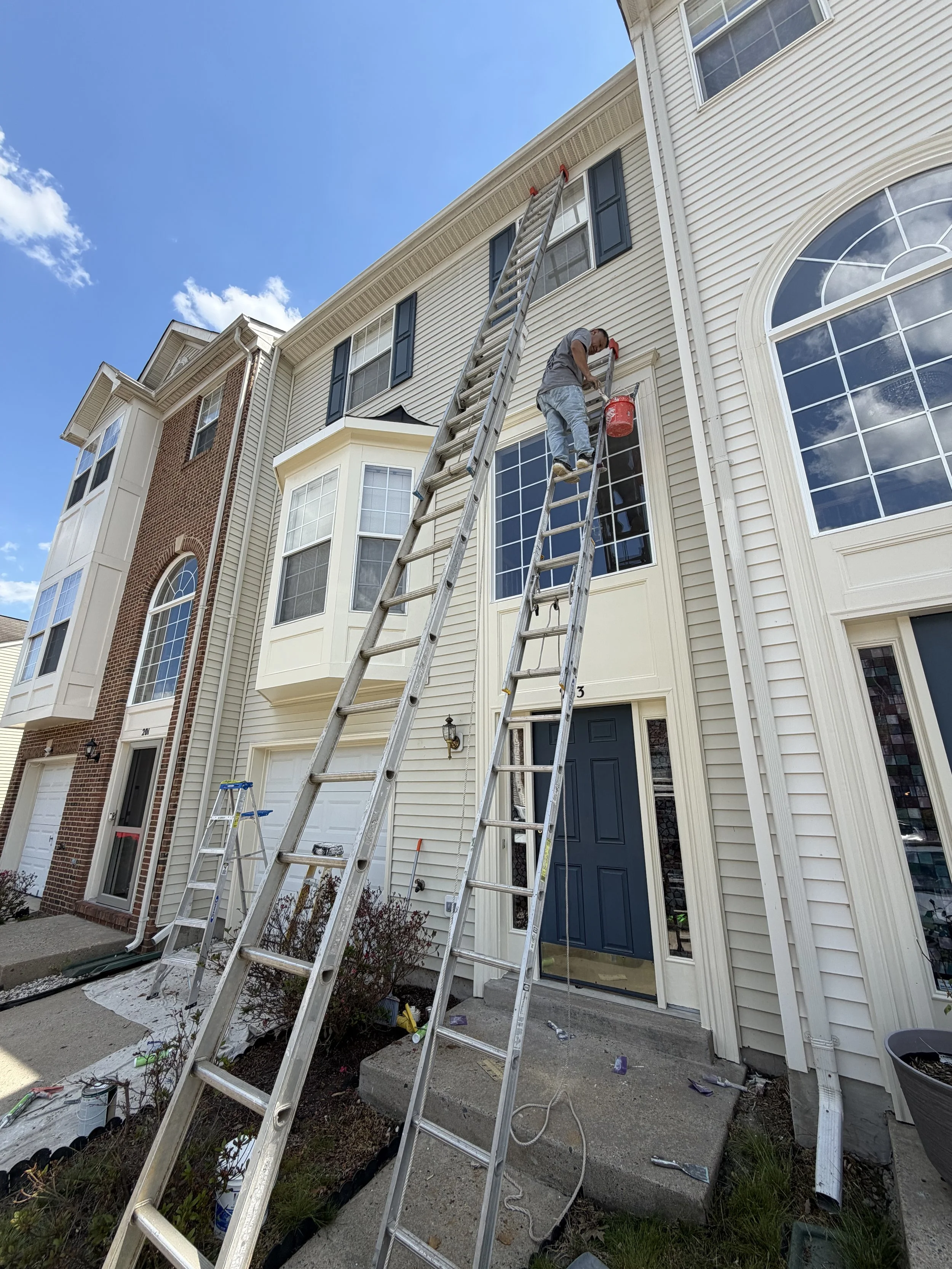 A man standing on a small ladder on the front porch of a multi-story house, painting or performing maintenance on the upper exterior wall, with two tall ladders leaning against the house and various tools and supplies on the ground.
