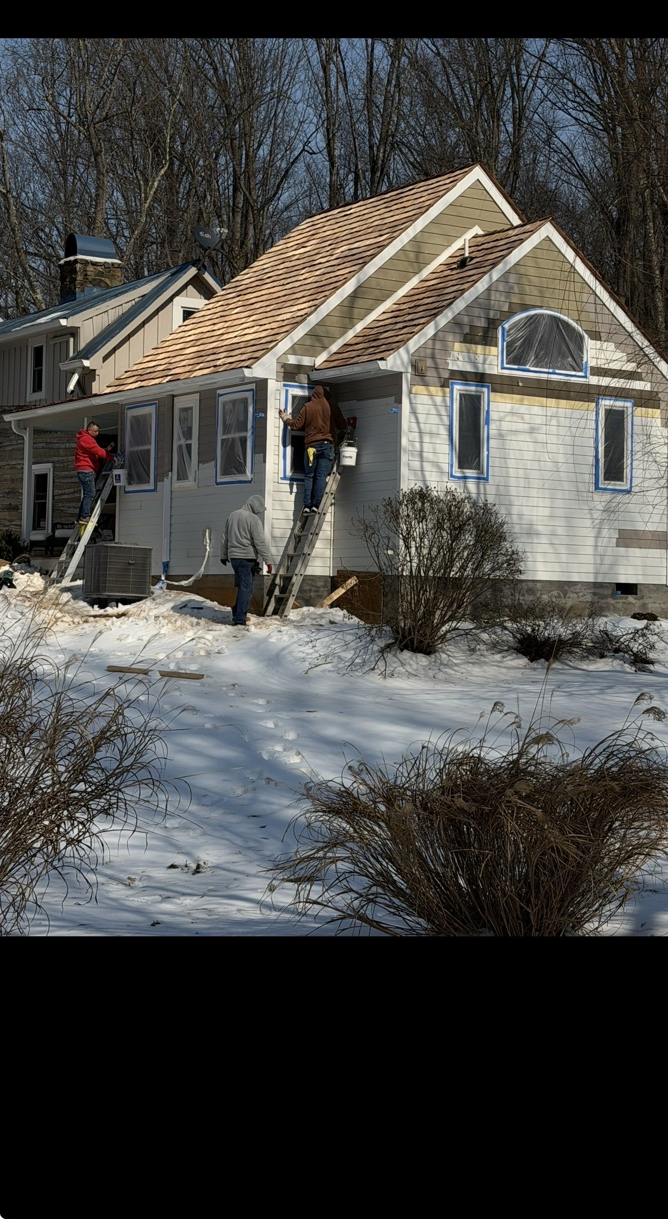 People working on the exterior of a house under construction during winter, with snow on the ground and leafless trees in the background.