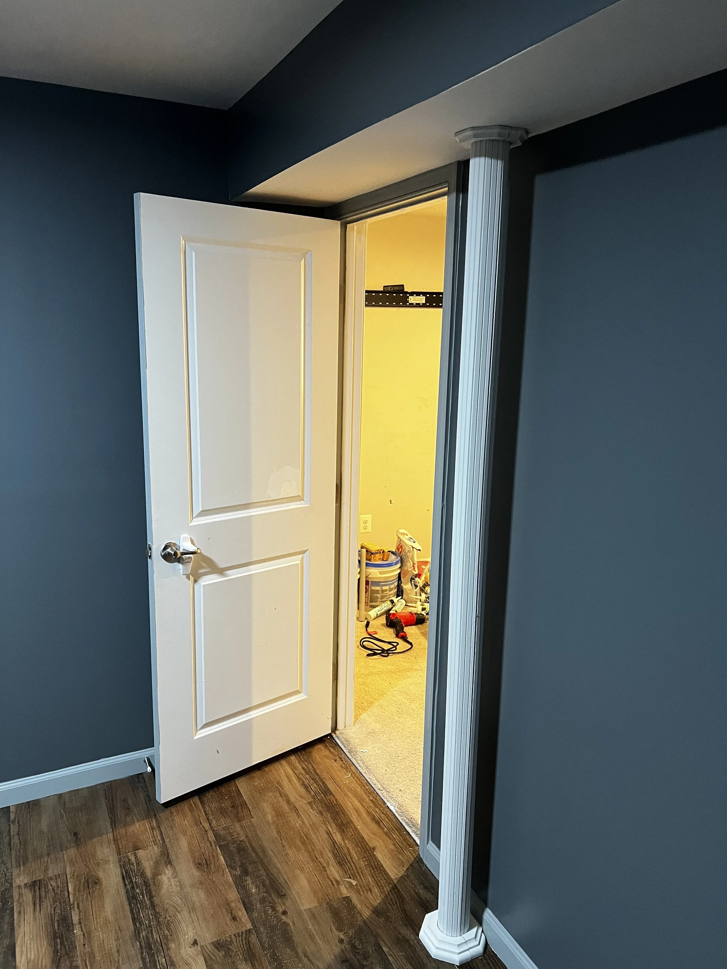 A partially open white door leading into a room with yellow walls and various tools and supplies on the floor, with a dark blue wall and a decorative column in the foreground.