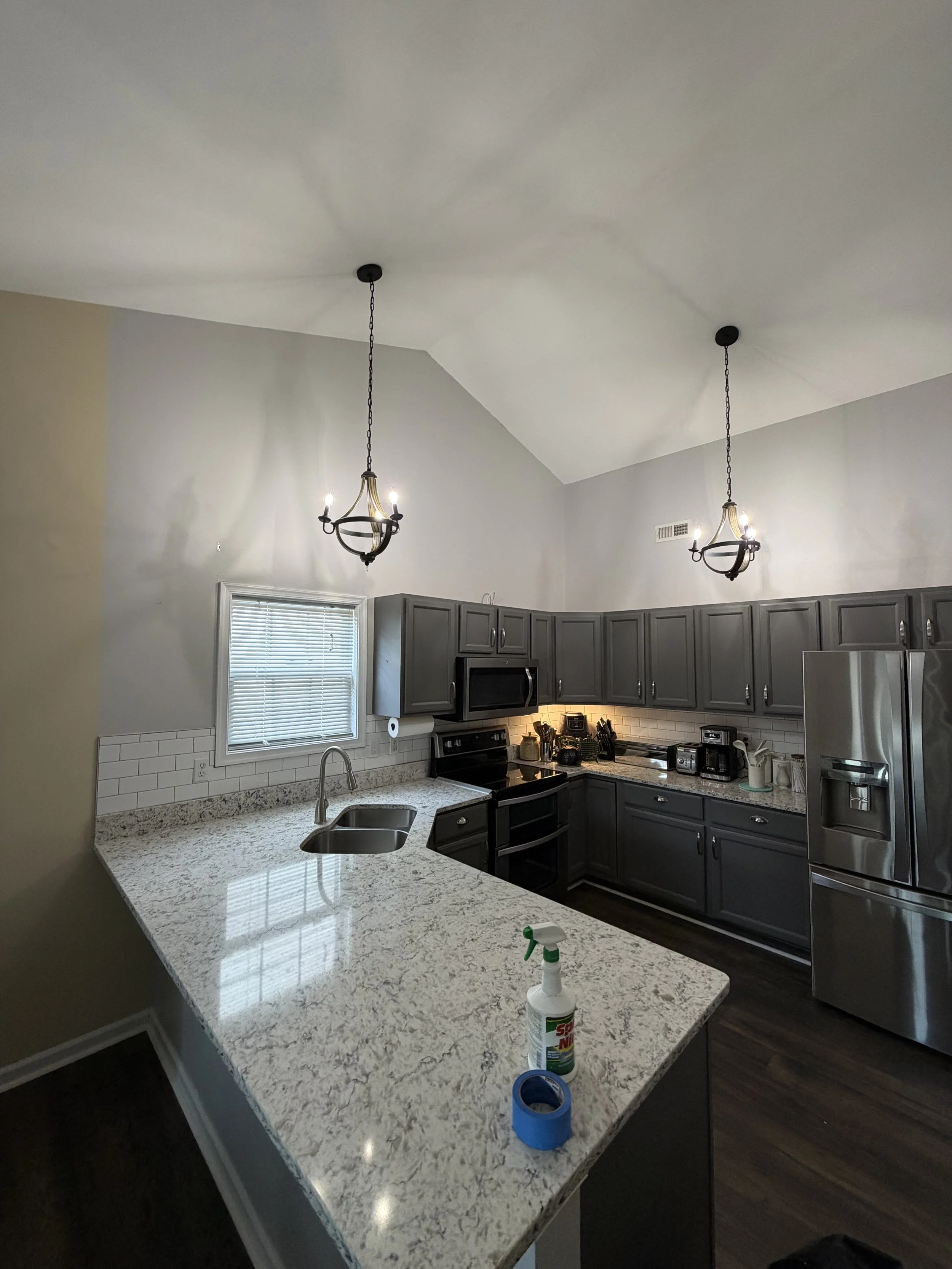 Kitchen with gray cabinets, stainless steel appliances, granite countertops, pendant lights, a window with blinds, a double sink, and cleaning supplies on the counter.