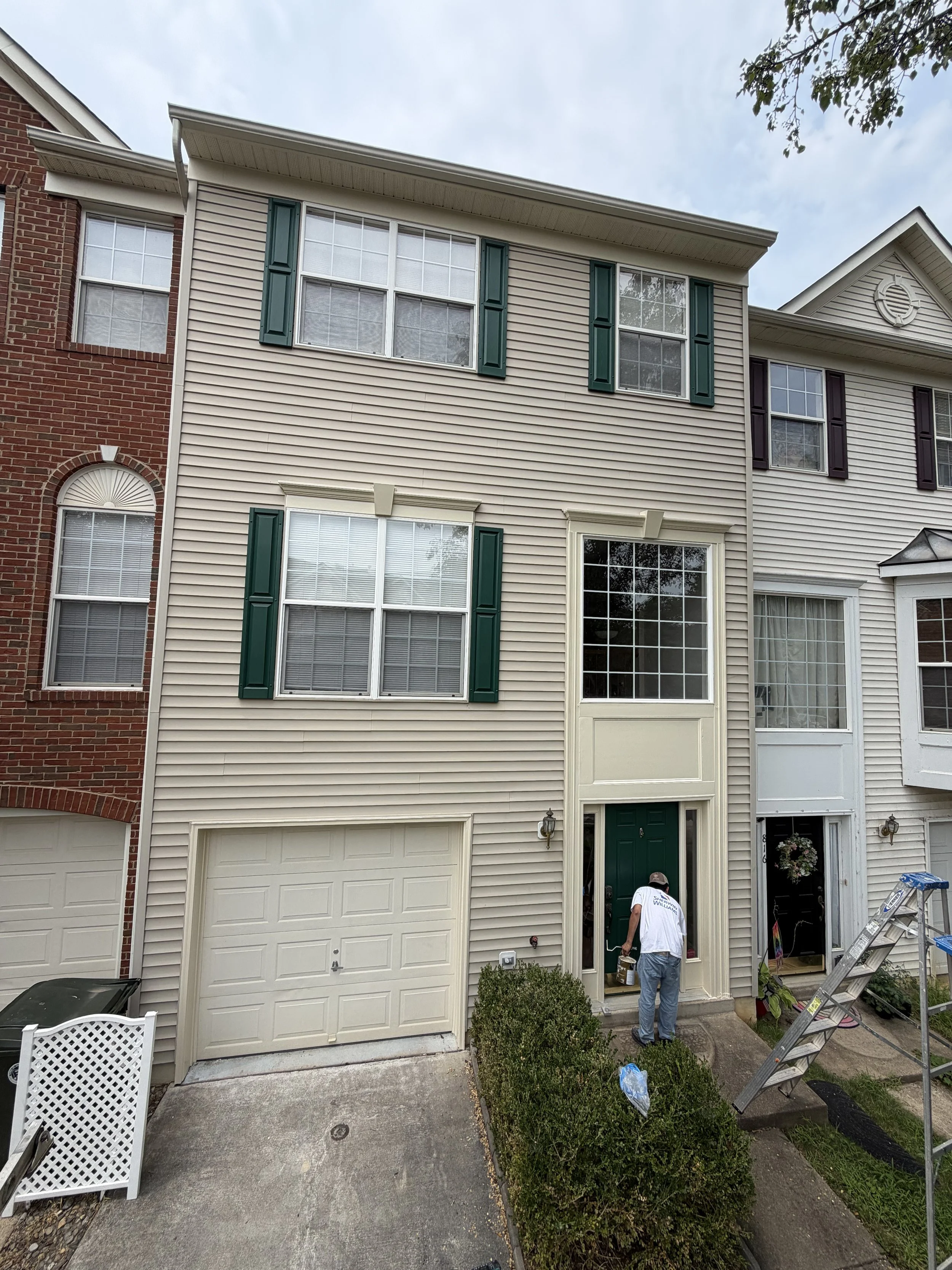 A person standing in front of a townhome with beige vinyl siding, green shutters, and a green front door. The person is wearing a white t-shirt and a cap, showing the back. A ladder is set up nearby, and there are various tools and items around, indi