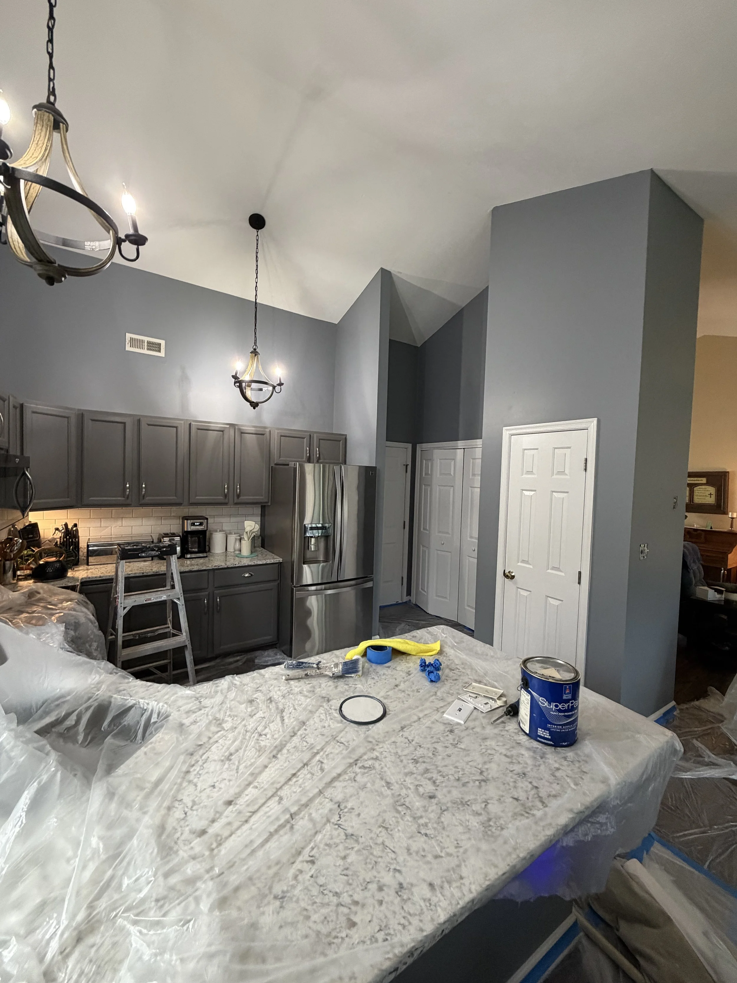 Kitchen under renovation with gray cabinets, two chandeliers, and a large white marble countertop covered with plastic, painter's tape, and painting supplies.