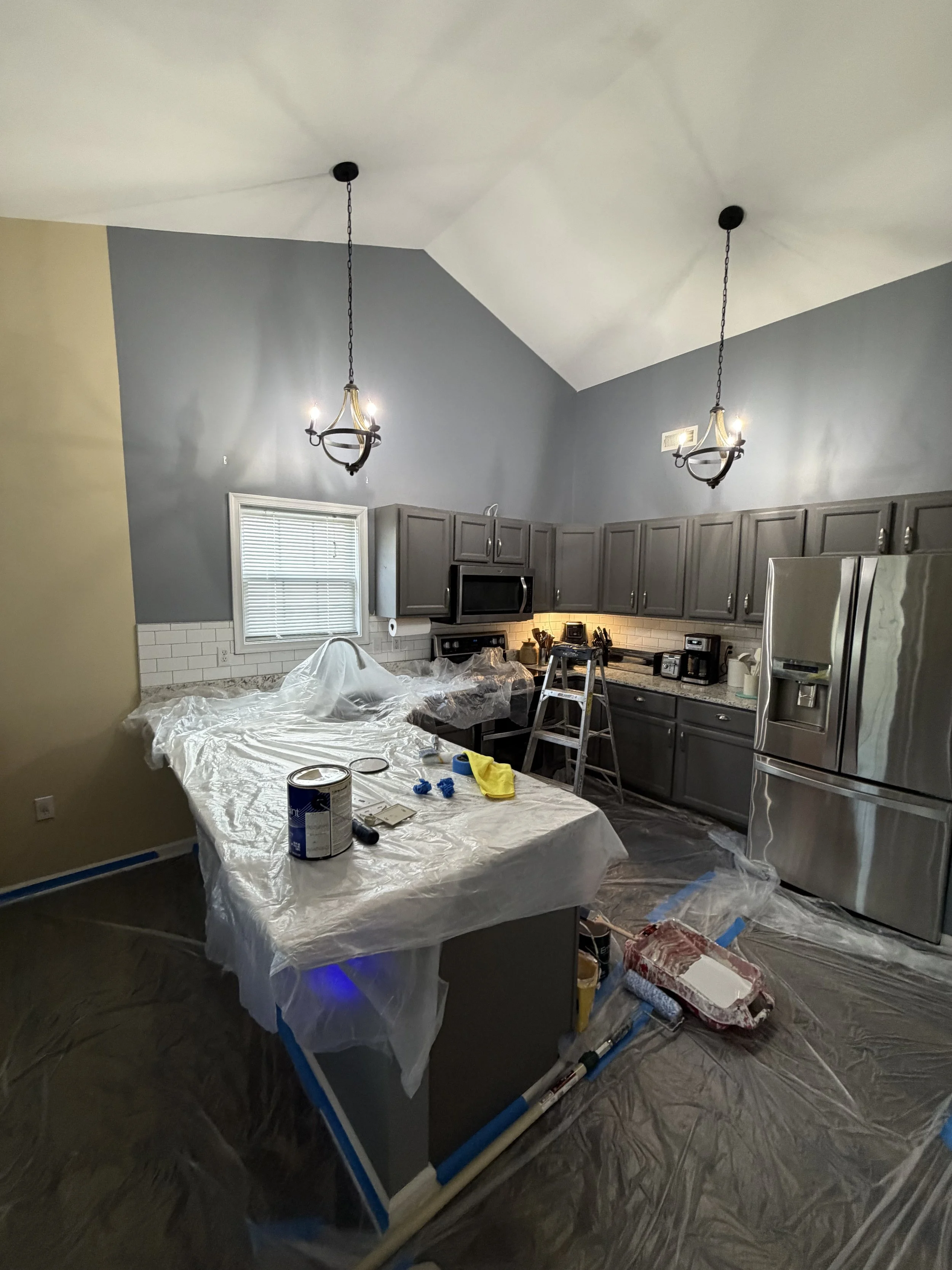 Kitchen undergoing renovation with painting supplies on a covered table, installed gray cabinets, new stainless steel appliances, and protective plastic sheets on the floor.