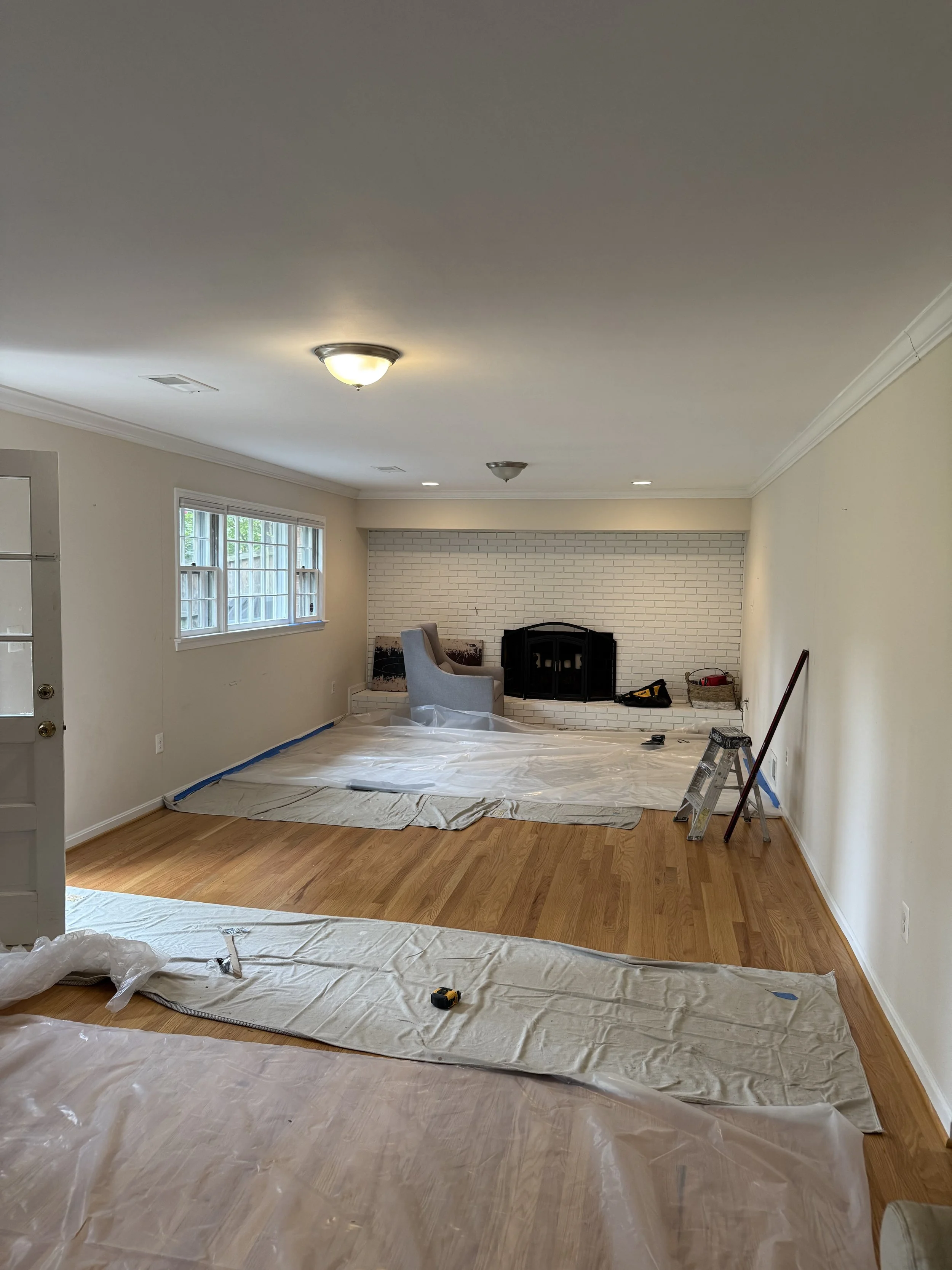 Living room undergoing renovation with a fireplace, two chairs, large window, and plastic sheets covering the hardwood floor.