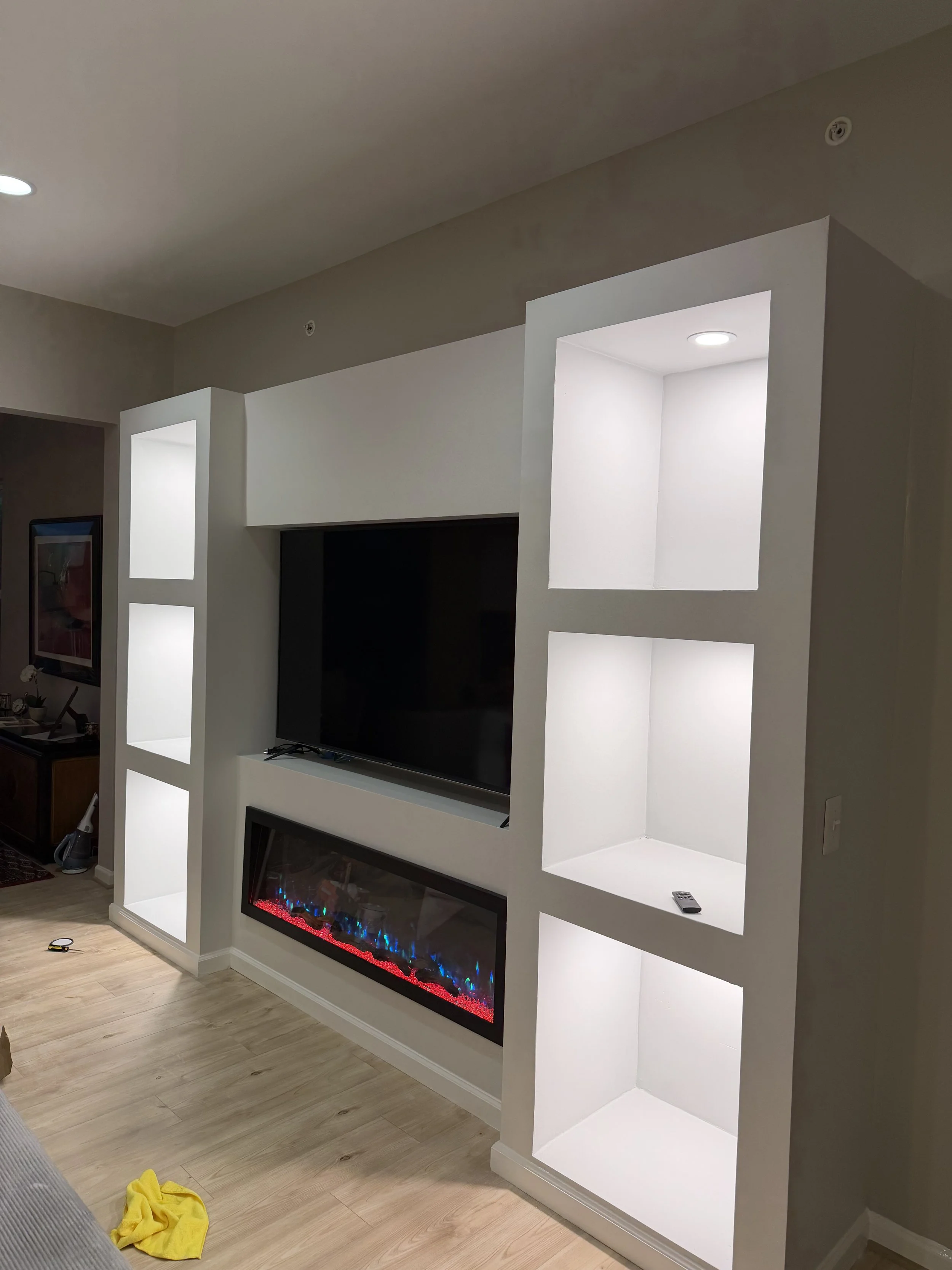 Living room wall with built-in illuminated display shelves, a flat-screen TV, and an electric fireplace with blue and red lighting.