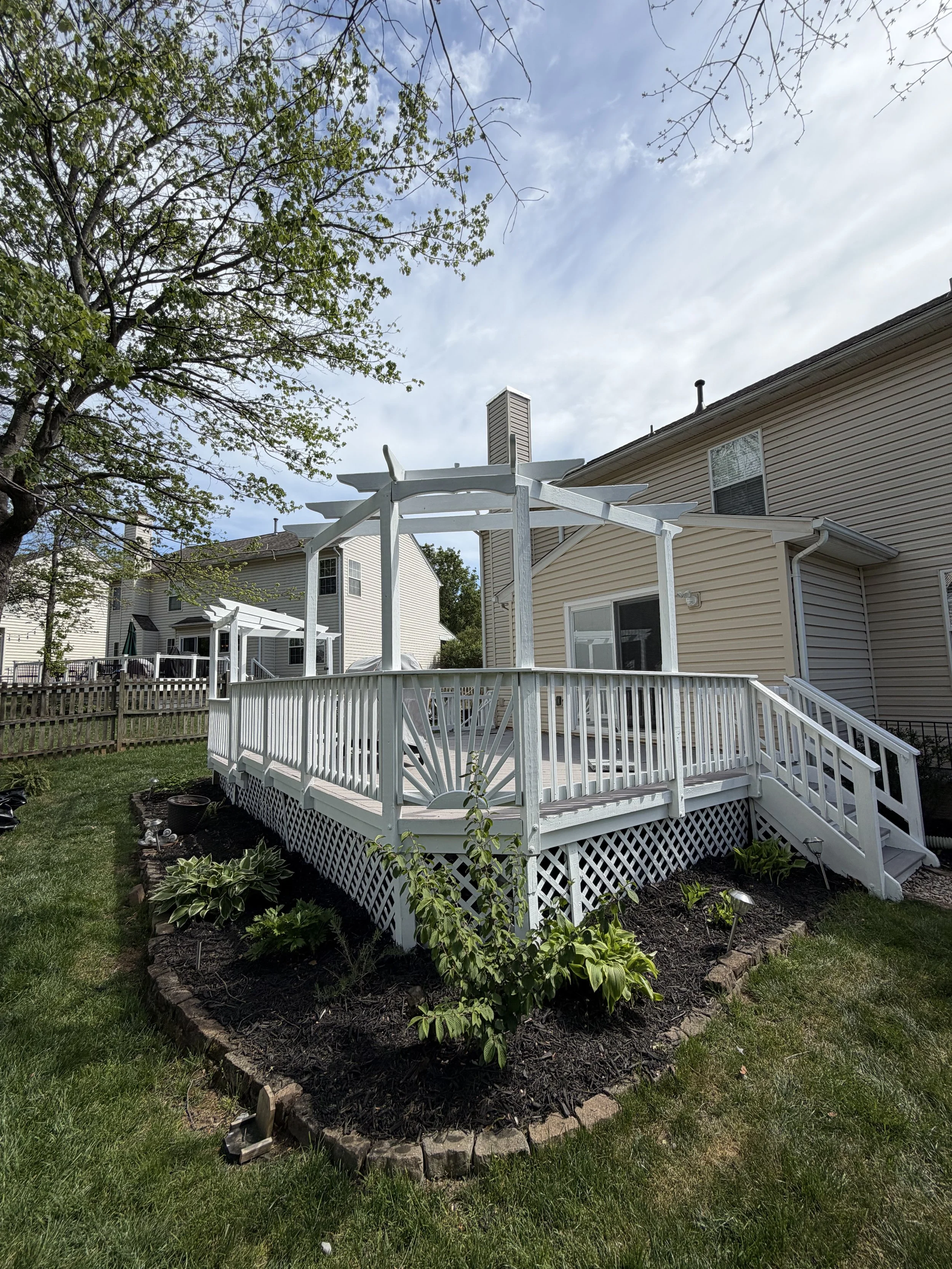 Backyard with white wooden deck and railing, built around a landscaped garden with plants and mulch, attached to a beige house with siding and a chimney, green grass, trees with leaves and bare branches, cloudy sky.