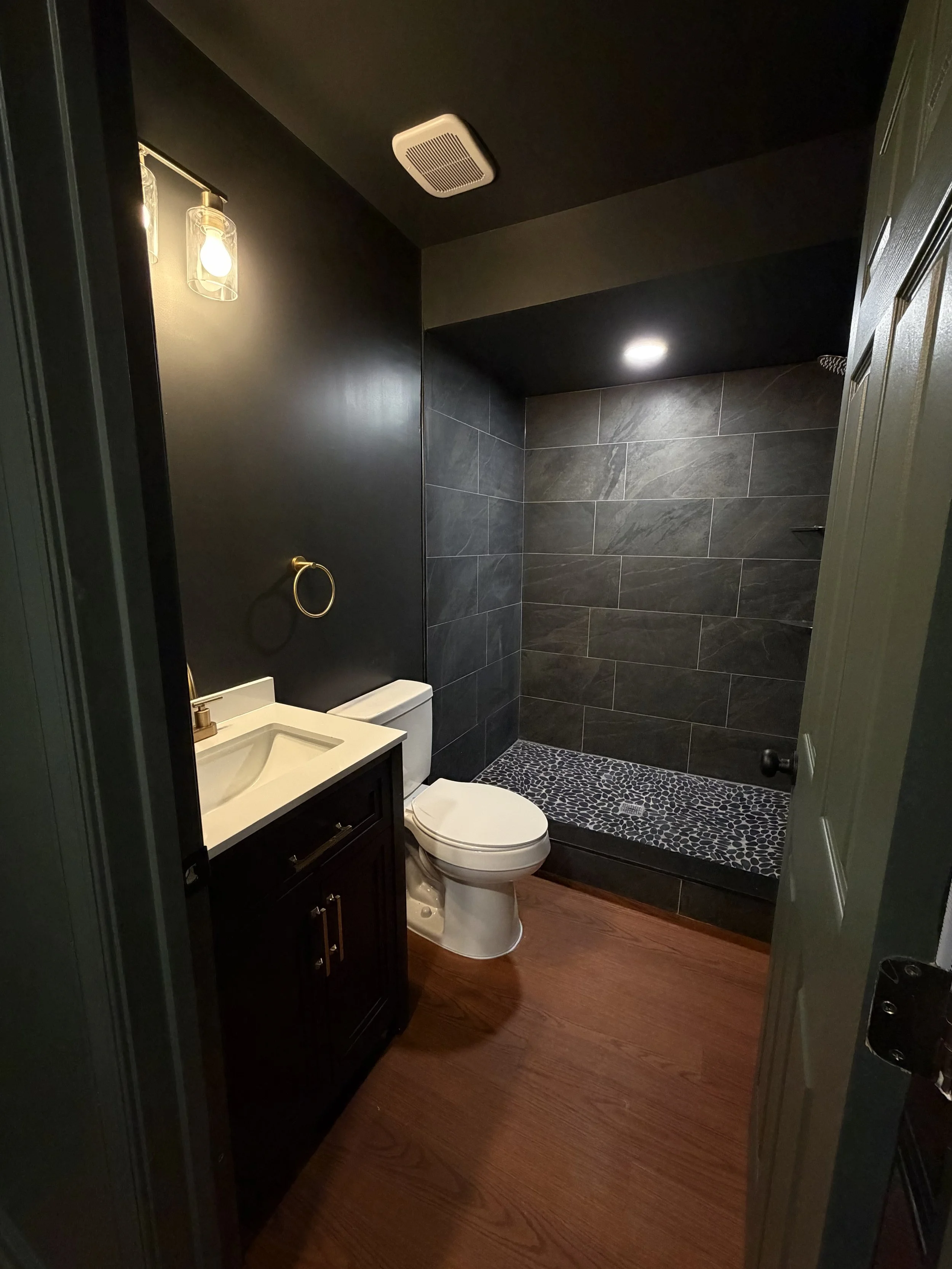 A small bathroom with black walls, a white toilet, a wooden vanity with a white sink, a gold towel ring, a patterned shower mat, and grey tiled shower area with a ceiling vent.