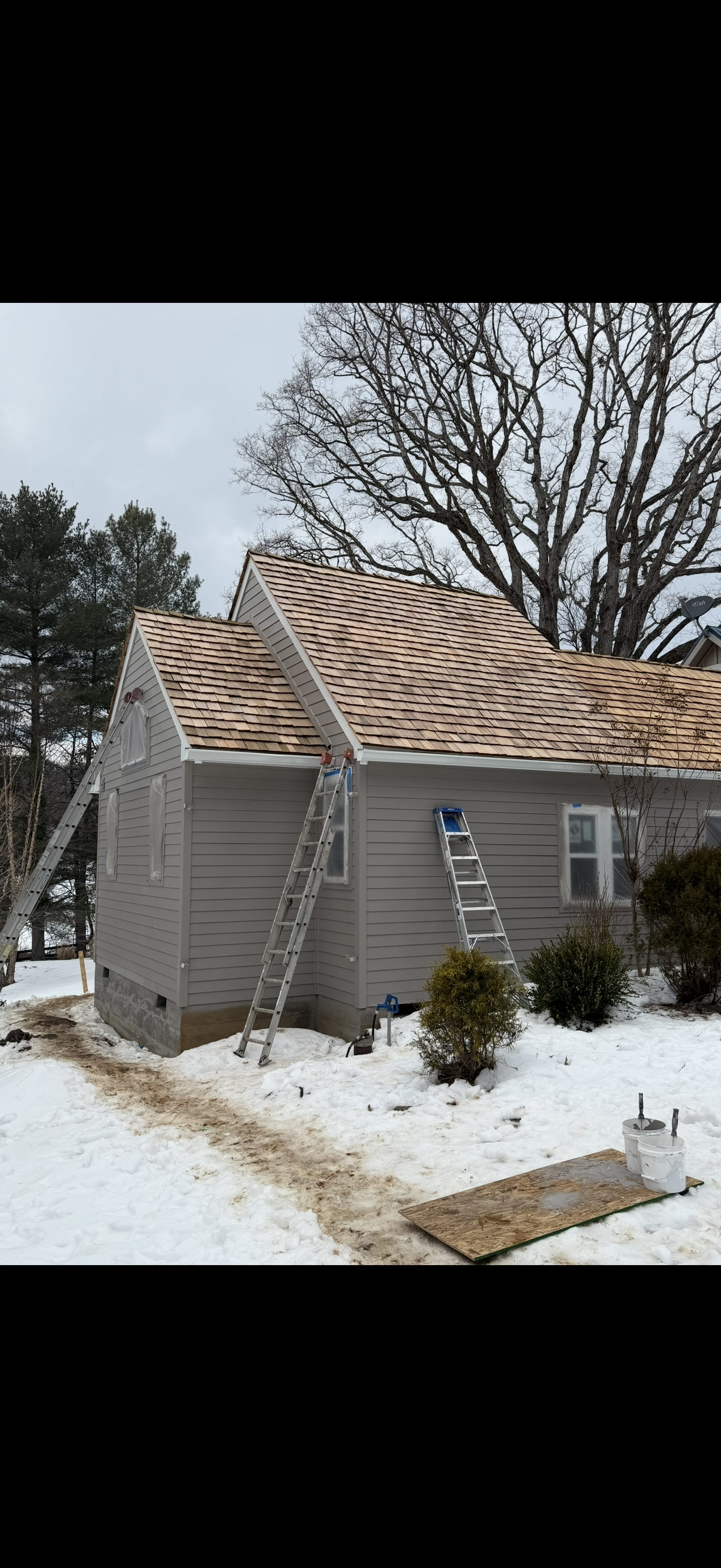 A house under construction with three ladders leaning against the roof, which has new shingles. Snow covers the ground, and there are trees in the background.