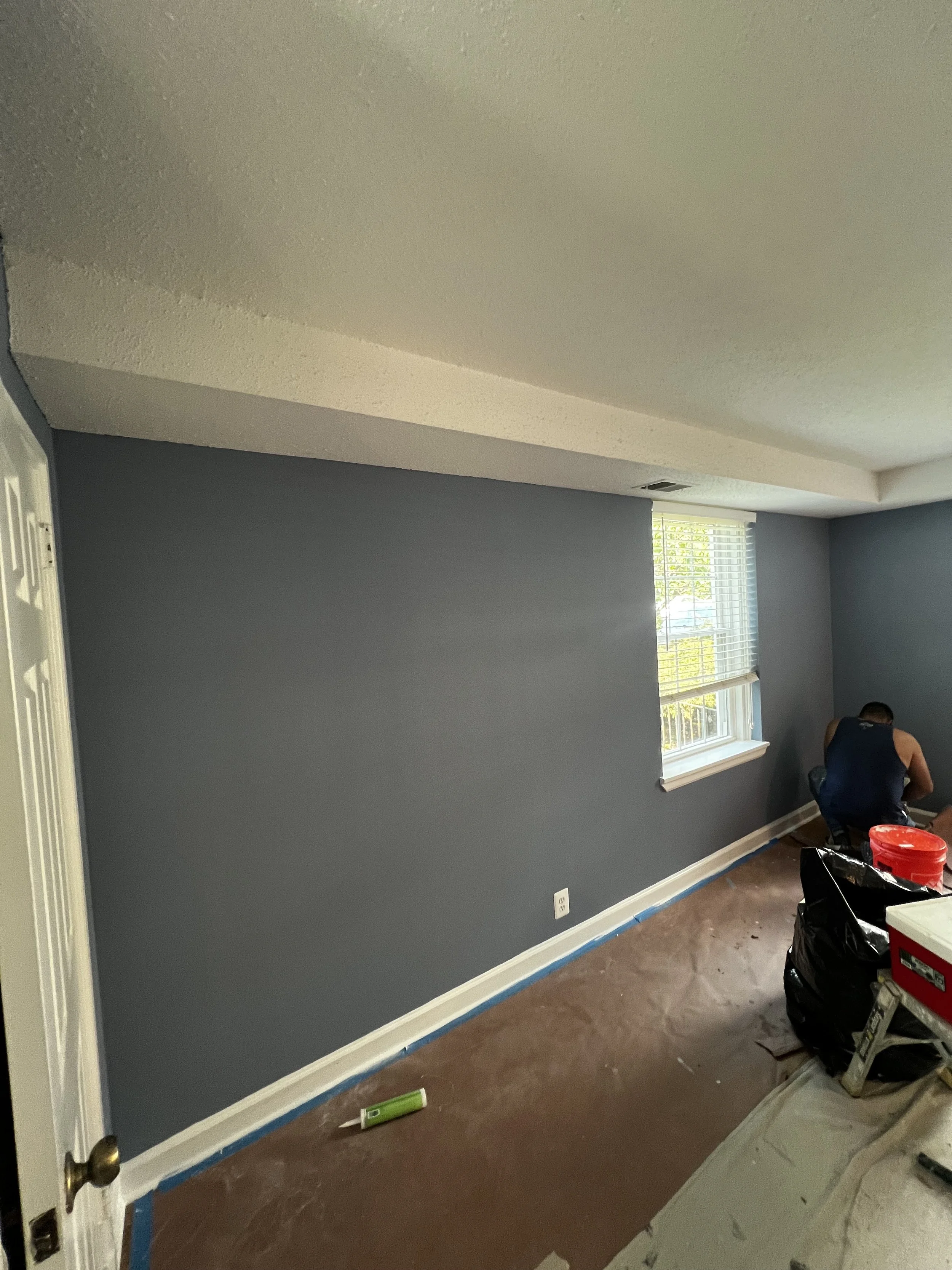 A room with blue painted walls and a white ceiling with textured finish. There are two windows with white blinds allowing natural light. A person is kneeling near the window, seemingly working on the wall. Painting supplies and tools are scattered on