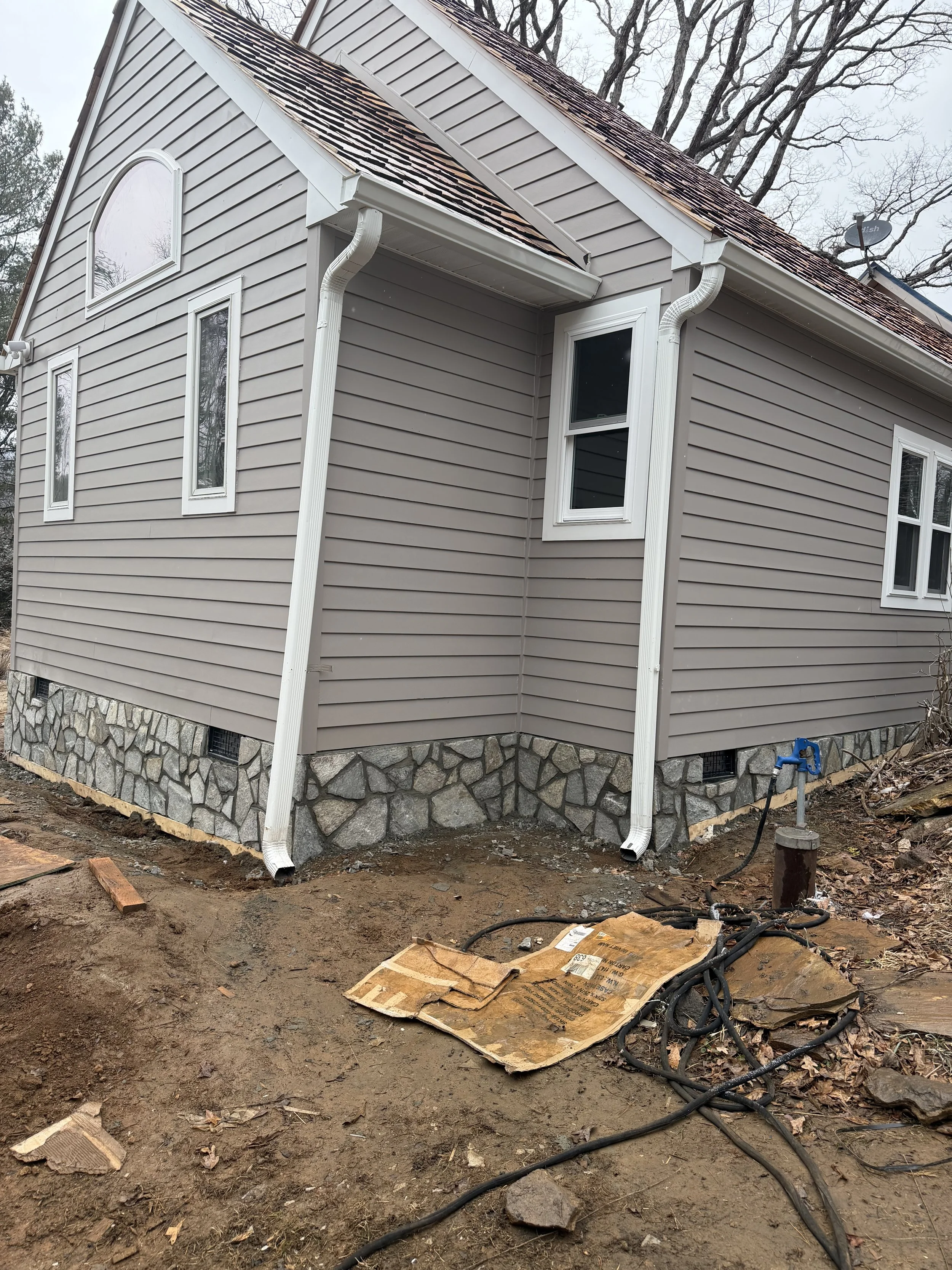 Exterior of a house under construction with beige vinyl siding, stone foundation, and white trim. The ground around is dirt with construction wires and materials, and trees are visible in the background.