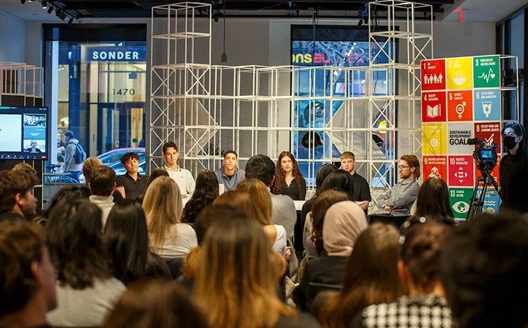 Panel discussion with seven people seated at the front of a room, audience listening, large display with UN Sustainable Development Goals, and a camera filming the event.