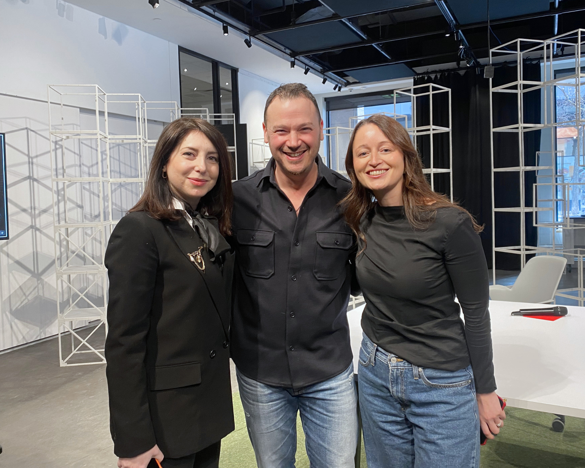 Three smiling people standing together indoors, with metal structures and large windows in the background.