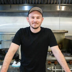 Portrait of a young man in a black t-shirt and gray cap, smiling, standing in a commercial kitchen.
