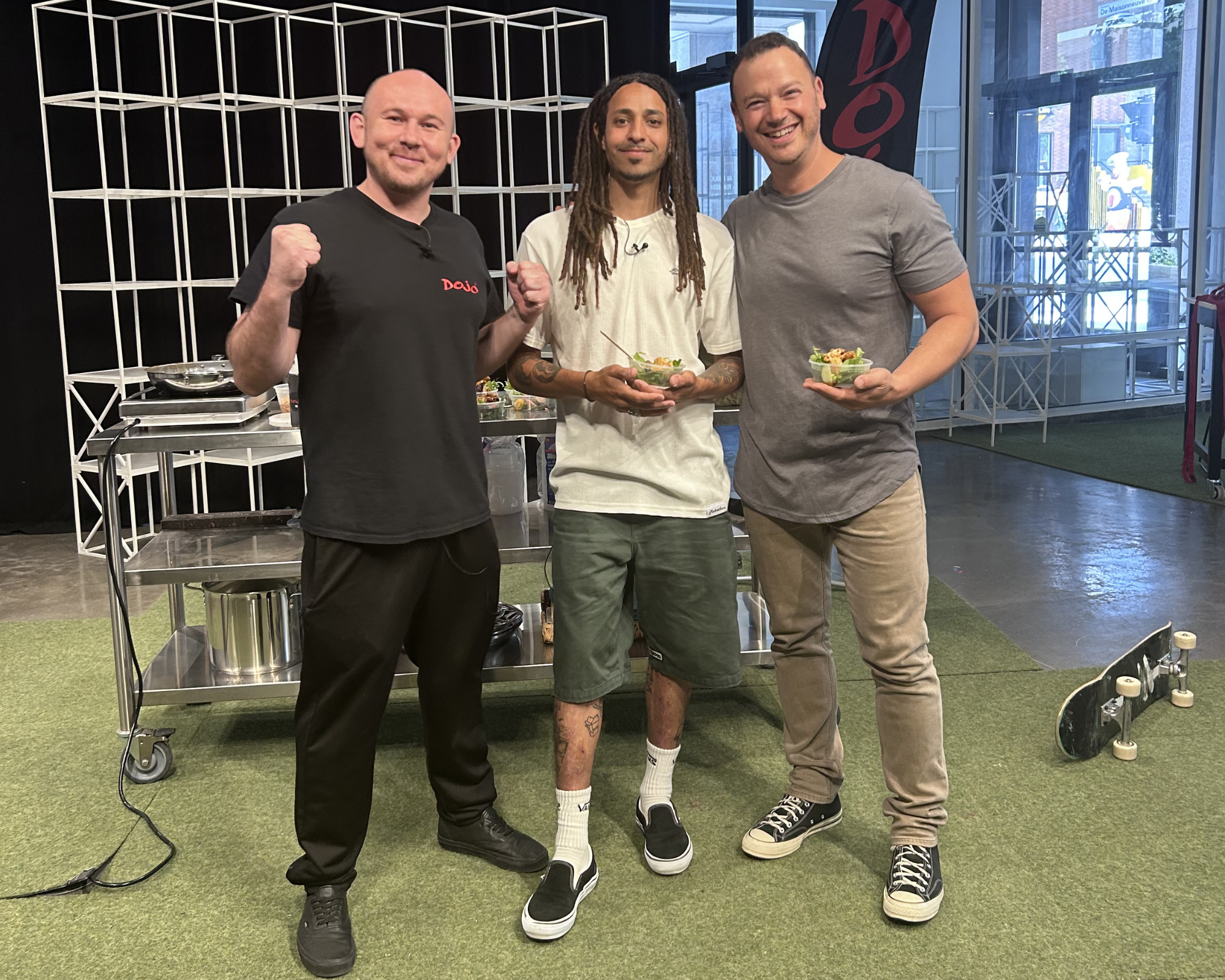 Three men standing indoors, smiling, holding bowls of salad, with one man making a fist gesture. The background features a black shelving unit, large windows, and a city street outside.