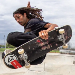 Skateboarder with dreadlocks performing a trick on a skateboard at an outdoor skate park.