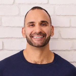 A smiling man with a beard and short hair wearing a navy blue shirt, standing in front of a white brick wall.